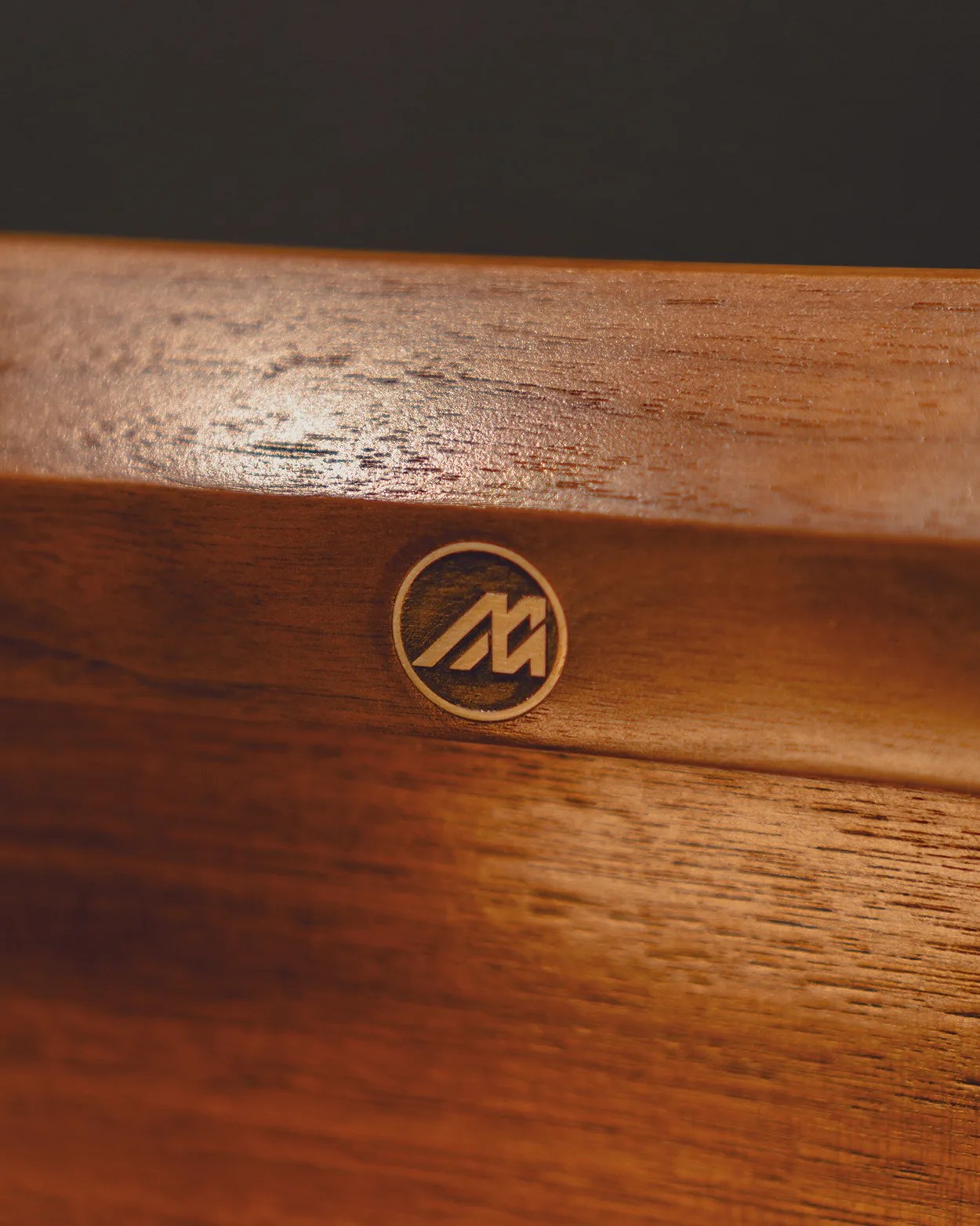 Close-up of a wooden surface with a circular logo engraved or inlaid, featuring stylized letters "M" and "A" inside the circle. The wood has a smooth, polished finish with visible grain patterns.