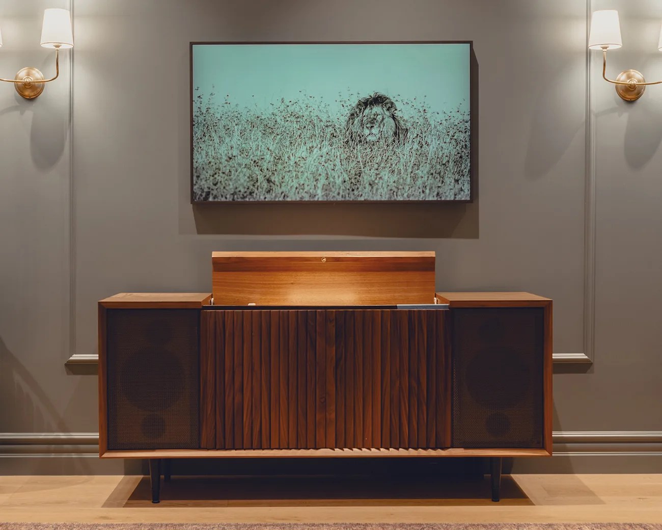 Mid-century modern wooden stereo console with a slatted front panel and two large speaker sections on either side, set against a gray wall. Above it hangs a framed picture of a lion partially hidden in tall grass. Two wall sconces with white lampshades flank the picture on either side. The floor is light wood with a rug partially visible at the bottom.