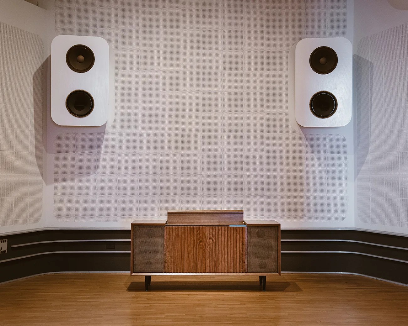 Mid-century modern wooden stereo console with built-in speakers, positioned against a wall with two large white wall-mounted speakers above it. The room has light-colored acoustic panels on the walls and a wooden floor.