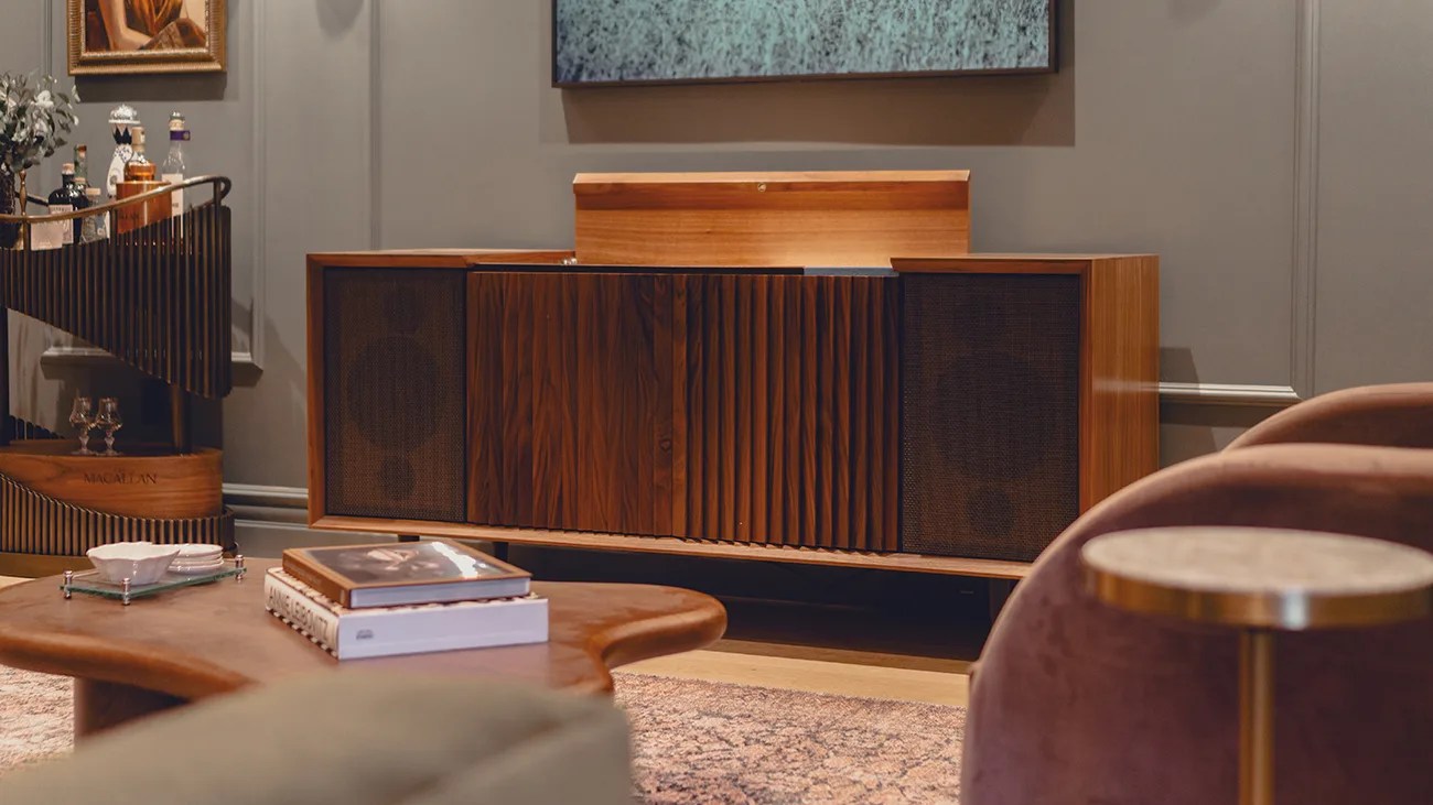 Wooden mid-century modern sideboard with a textured front panel and two speaker grills on either side, placed against a gray wall. A wooden coffee table with books and a small dish is in the foreground, and a spiral wooden bar cart with bottles is visible to the left. A round side table with a marble top and brass base is partially visible on the right.