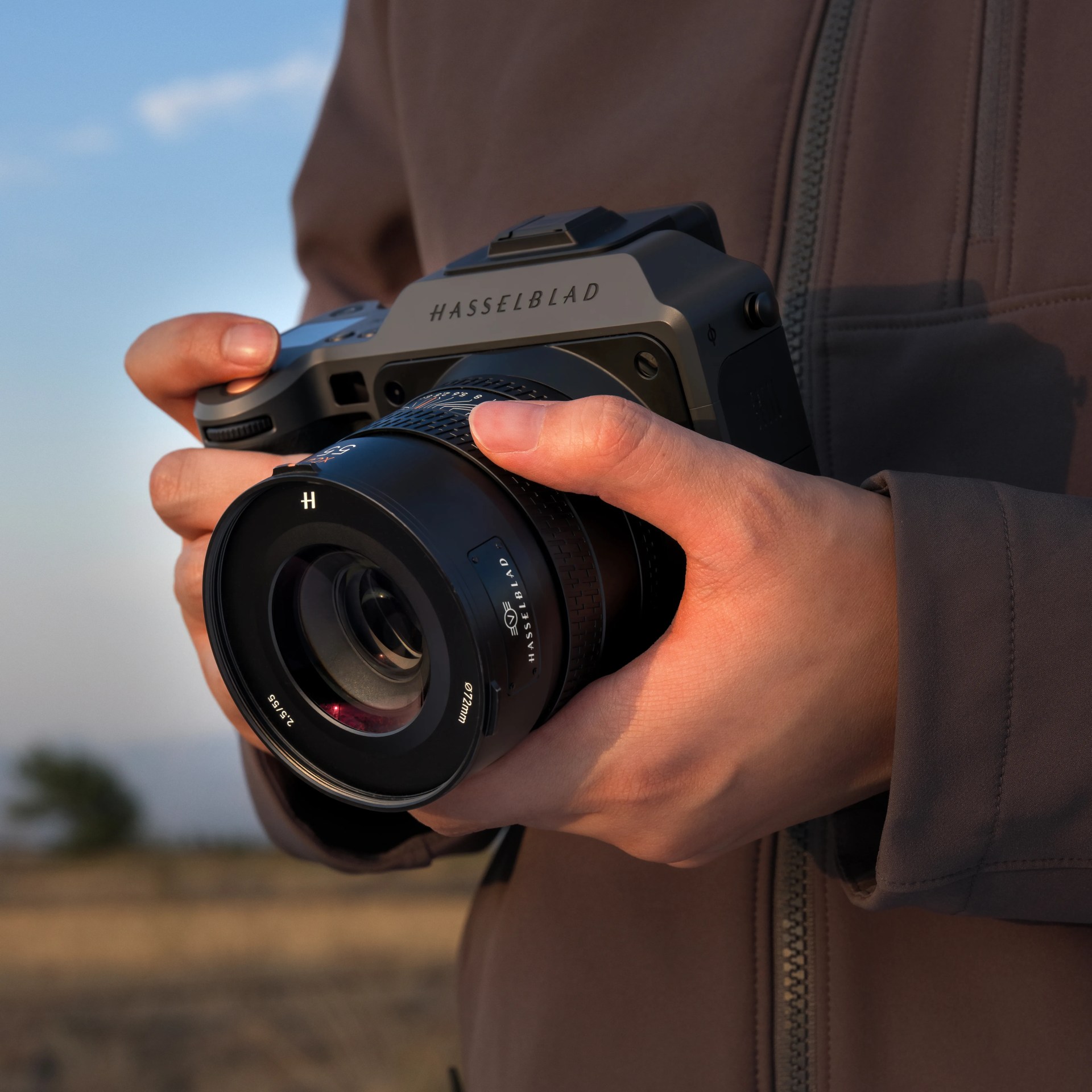 Hands holding a Hasselblad camera with a large lens, outdoors during golden hour with a blurred natural background. The person is wearing a brown jacket with a visible zipper.