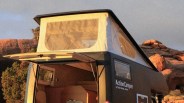 A black camper van with a raised white pop-up roof featuring mesh windows. The rear door is open, revealing a wooden interior with seating. The side of the camper has the text "ActionCamper by Trailer Design, Austria." The background includes rocky terrain and some greenery.