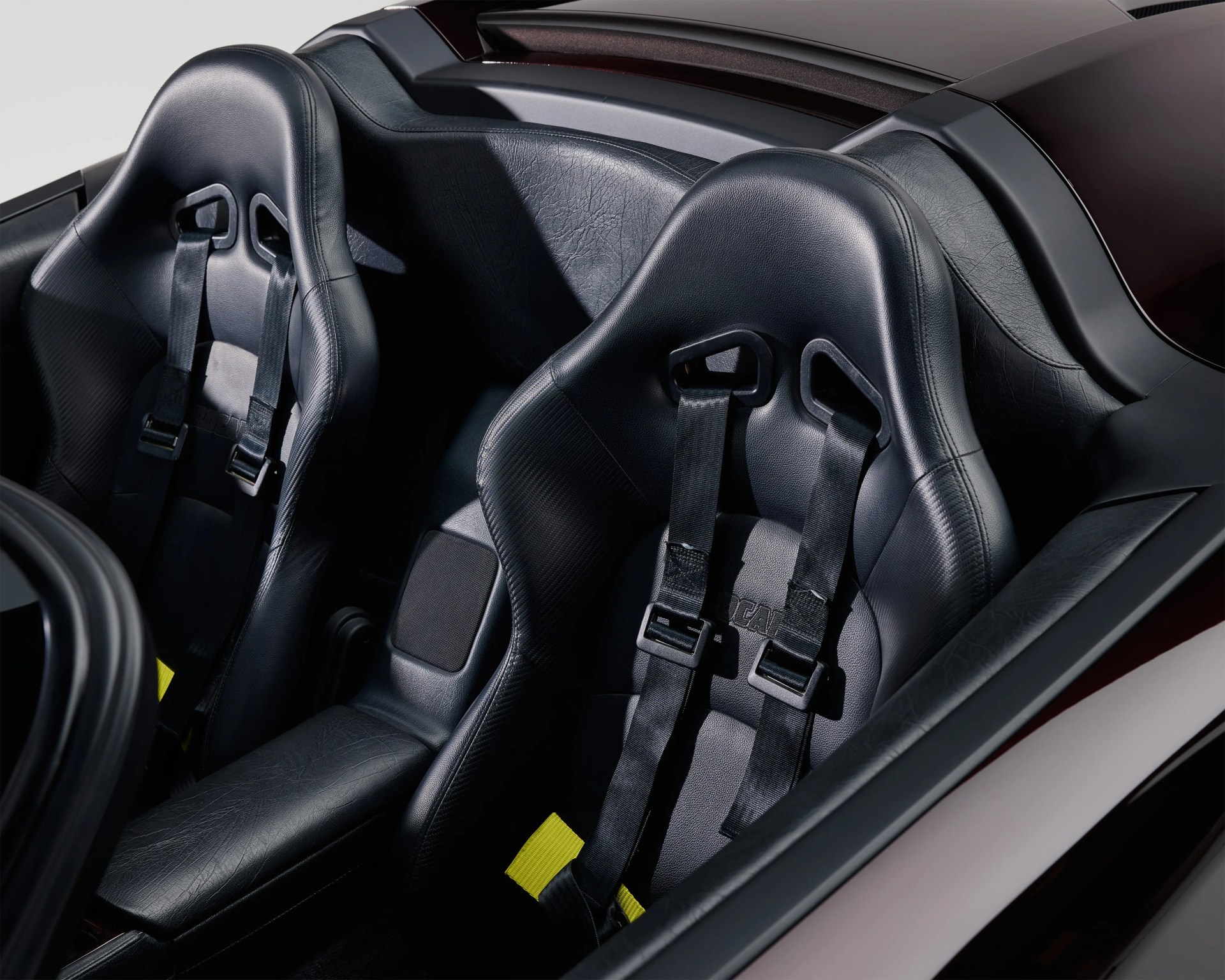 Two black racing bucket seats with integrated four-point harnesses, featuring leather upholstery and carbon fiber texture accents, inside a sports car. The seats have high side bolsters for support and yellow tags on the harness straps.