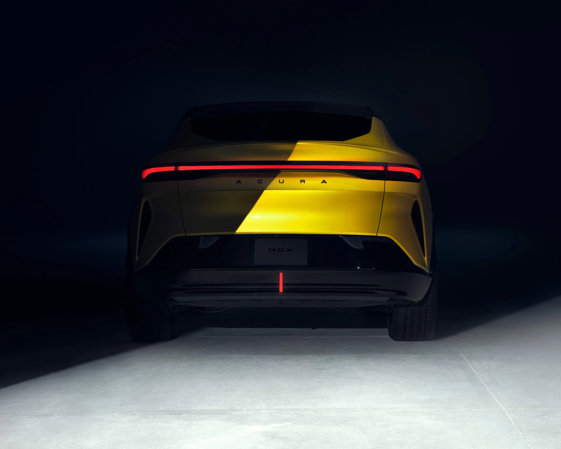 Rear view of a yellow Acura RSX prototype car with a continuous red taillight strip across the back, set against a dark background with a spotlight illuminating the vehicle.