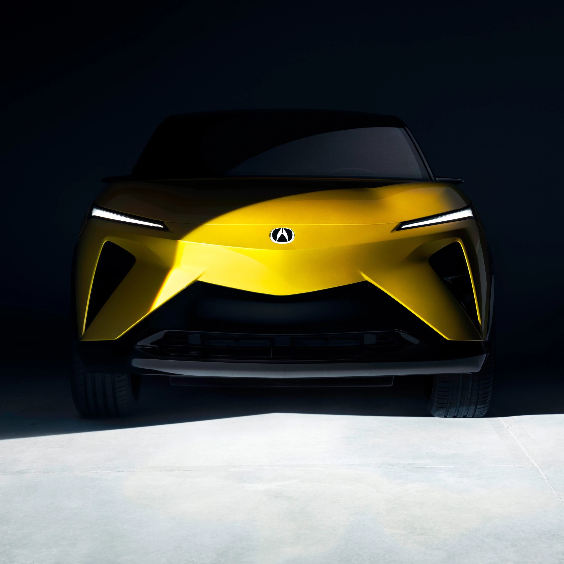 Front view of a sleek, modern yellow car with sharp, angular design elements and slim horizontal LED headlights, set against a dark background with a spotlight highlighting the center of the hood and front bumper.