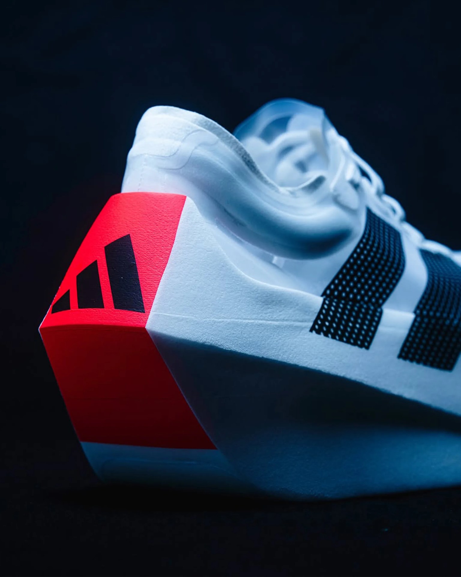 White athletic shoe with black stripes on the side and a distinctive angular red heel featuring a black Adidas logo, set against a dark background.