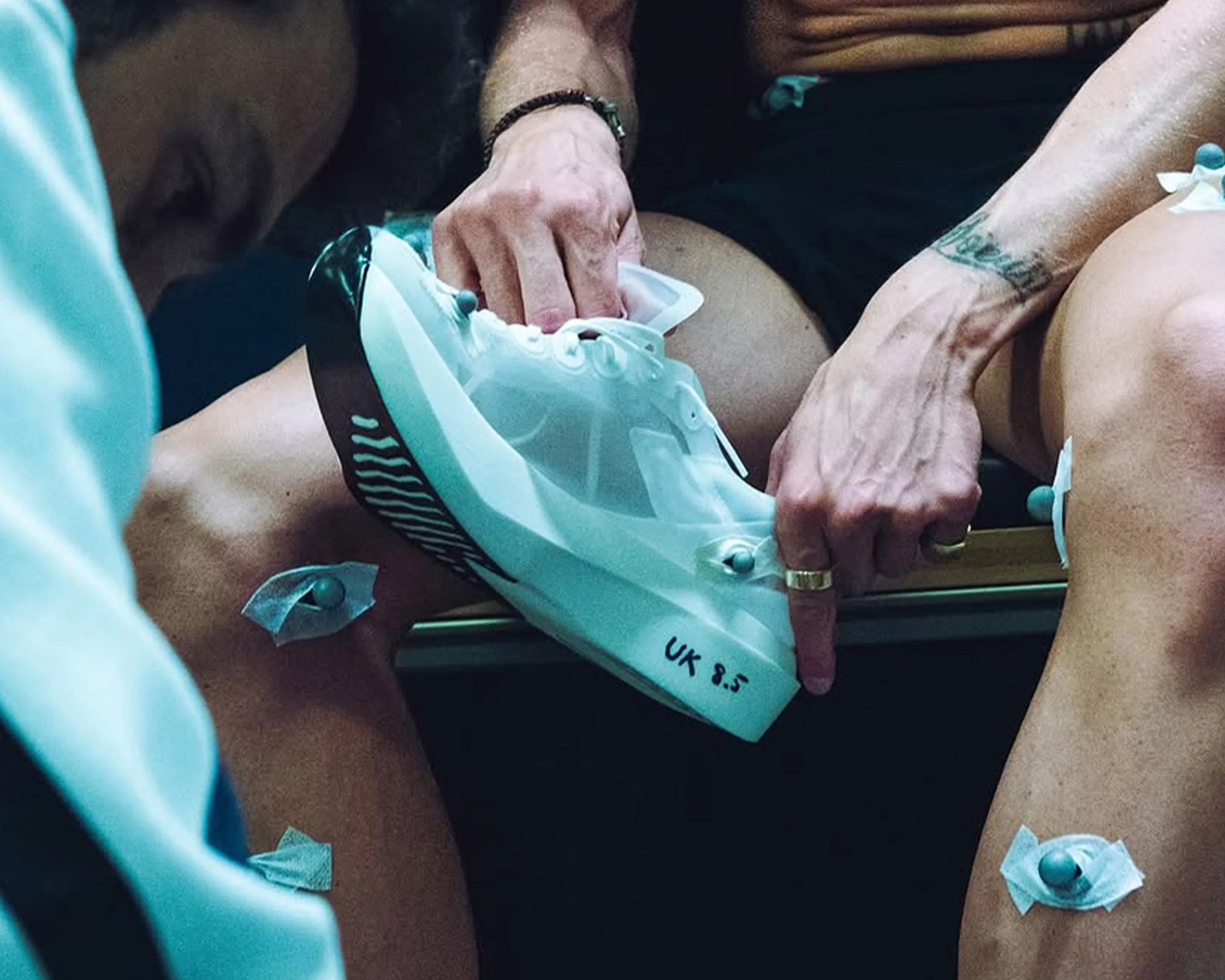 A person is holding a white athletic shoe marked with "UK 8.5" on the sole. The person has a tattoo on their forearm and is wearing rings and a bracelet. Several small round sensors are attached to their legs with tape, likely for motion capture or biomechanical analysis. The person is seated, wearing black shorts.