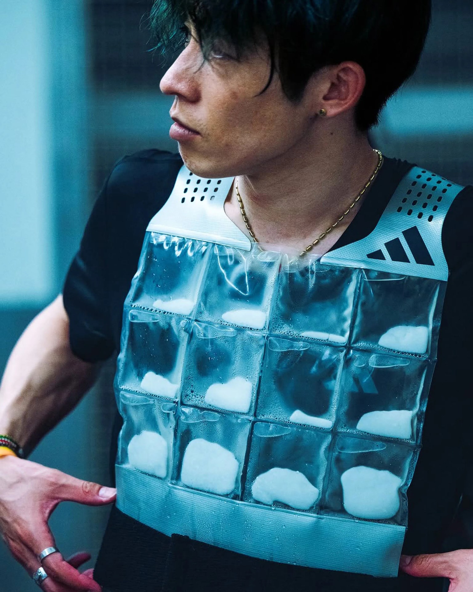 A person wearing a black shirt and a cooling vest with multiple transparent pockets filled with ice packs. The vest has white shoulder straps with black ventilation holes and a black logo on the right side. The person has short dark hair, a gold chain necklace, and is looking to the left.