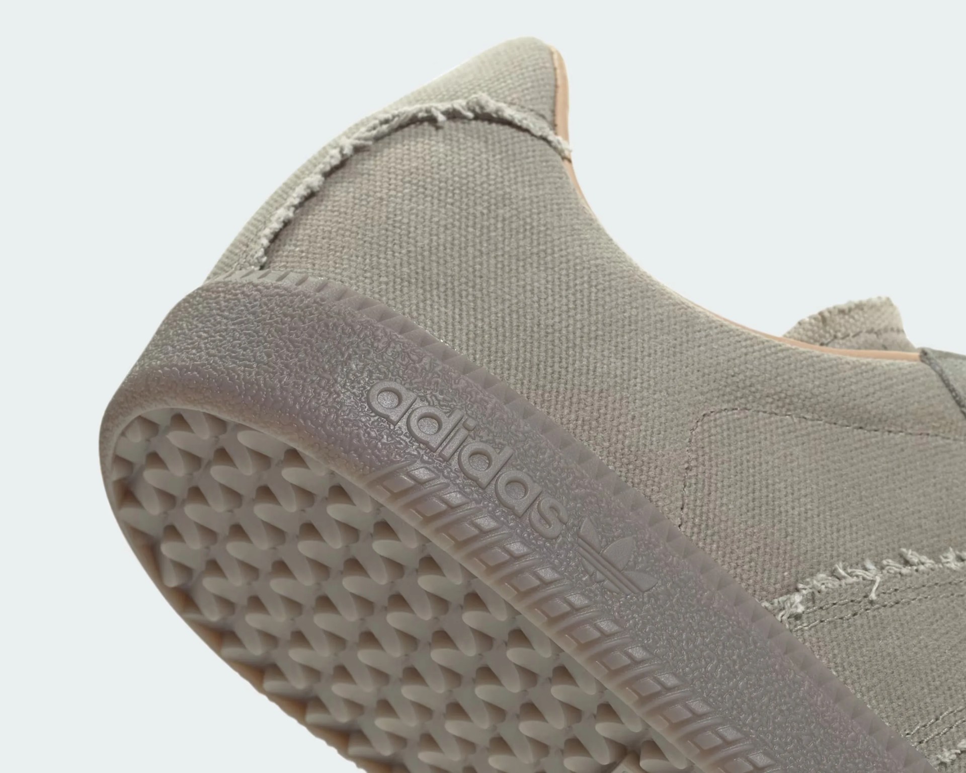 Close-up of the heel and sole of a light gray Adidas sneaker, featuring a textured rubber sole with a patterned tread and the Adidas logo embossed on the side. The upper part is made of a woven fabric with visible stitching and a slightly frayed seam detail.
