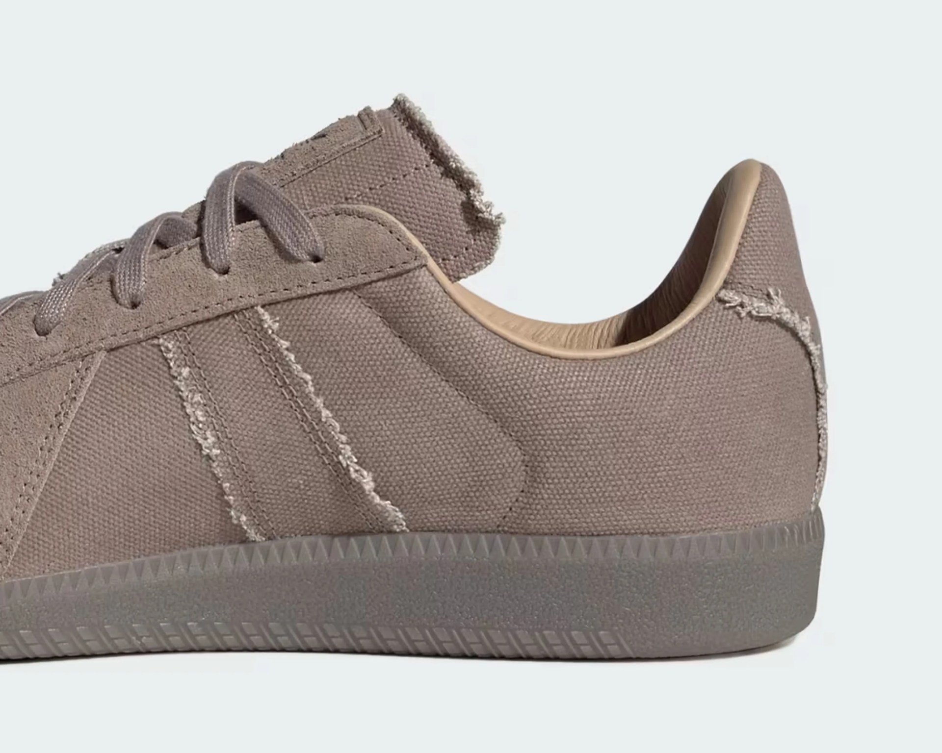 Close-up of the back and side portion of a taupe-colored sneaker with a textured fabric upper, frayed edges along the stripes and collar, and a matching rubber sole. The interior lining appears to be a smooth, light tan material. The sneaker features laces in a similar taupe shade.