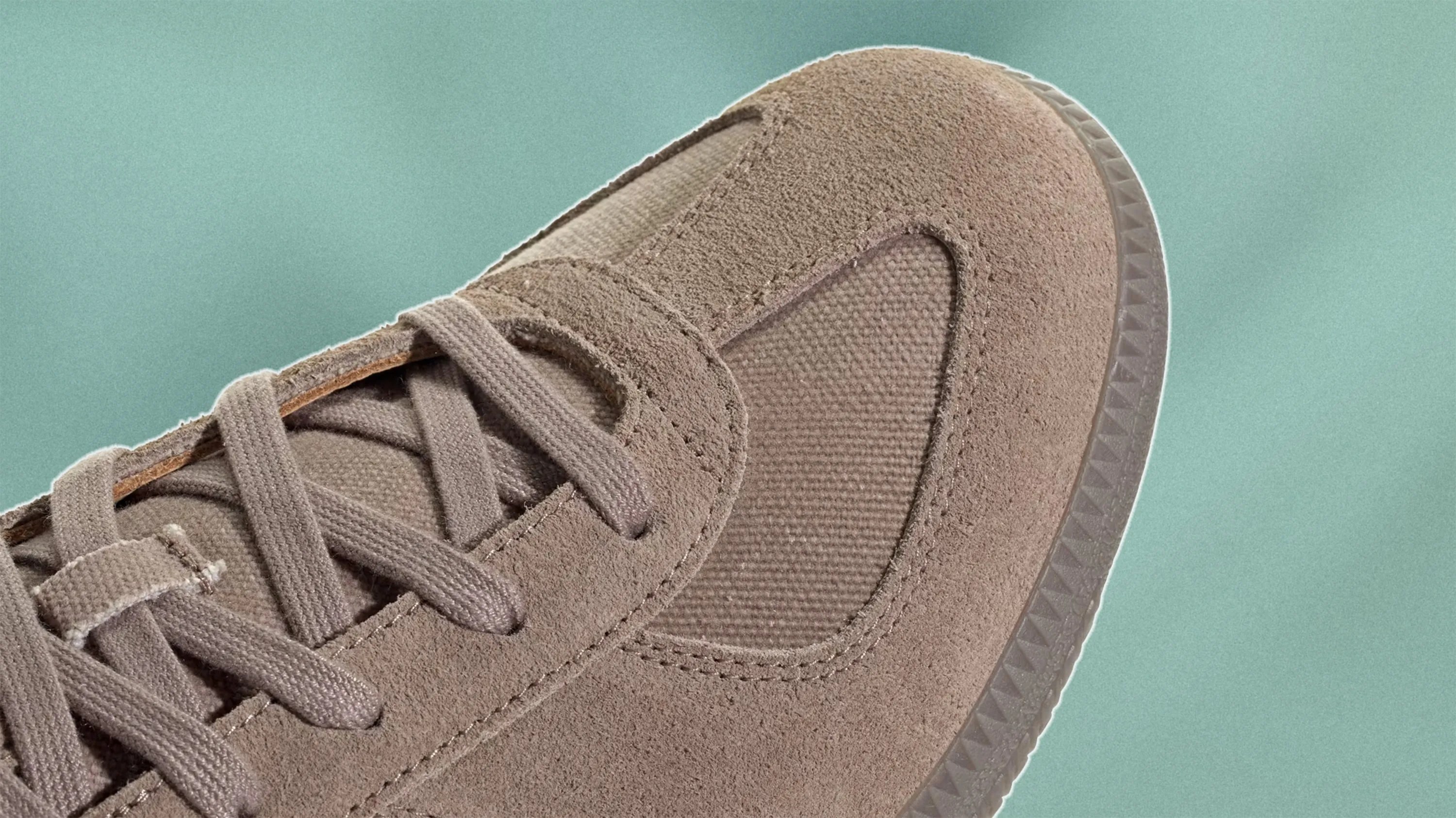 Close-up of a taupe-colored sneaker showing the laces, suede upper, and textured fabric panel near the toe, set against a muted green background.