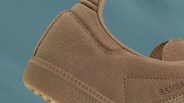 Close-up of the heel and side of a brown suede sneaker with a textured rubber sole and visible stitching. The shoe has a smooth leather trim around the ankle opening. The word "SAMBA" is partially visible on the side. The background is a solid muted blue color.