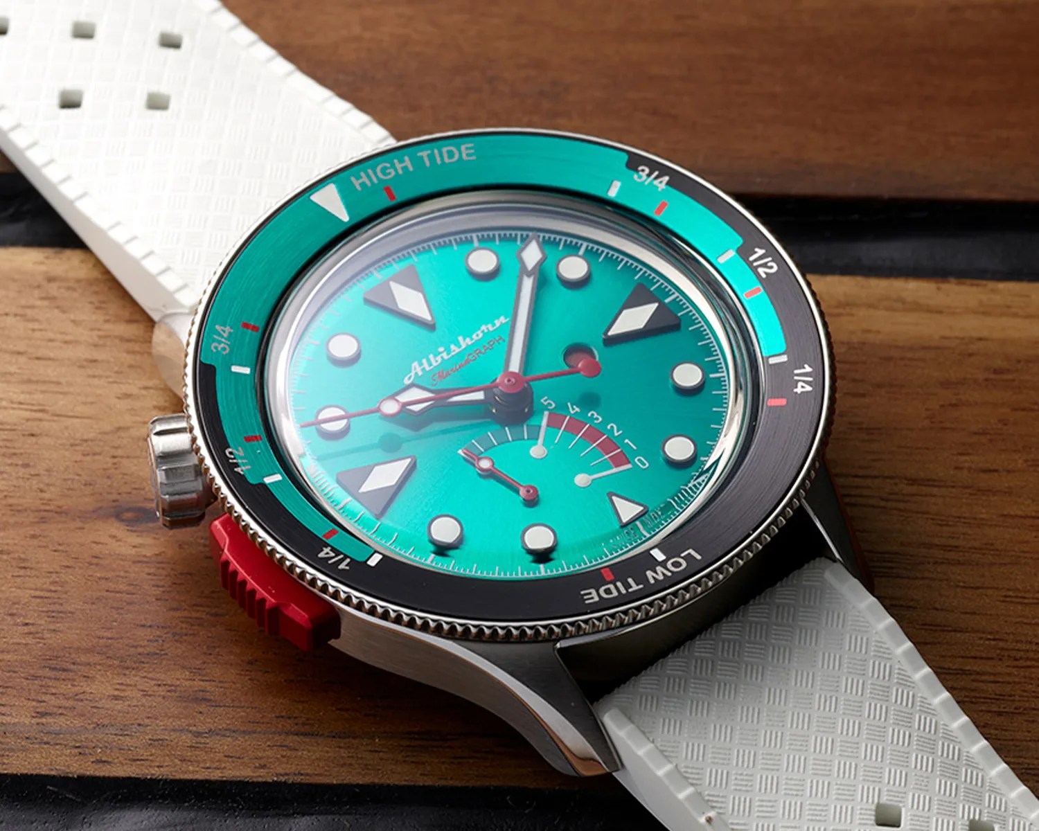 A wristwatch with a turquoise dial featuring large white and black geometric hour markers and a red second hand. The bezel is black and turquoise with markings for "HIGH TIDE" and "LOW TIDE." The watch has a white textured strap and a red crown on the side. The dial also includes a small gauge with numbers from 0 to 5 and two small hands. The brand name "Albishorn" is visible on the dial.