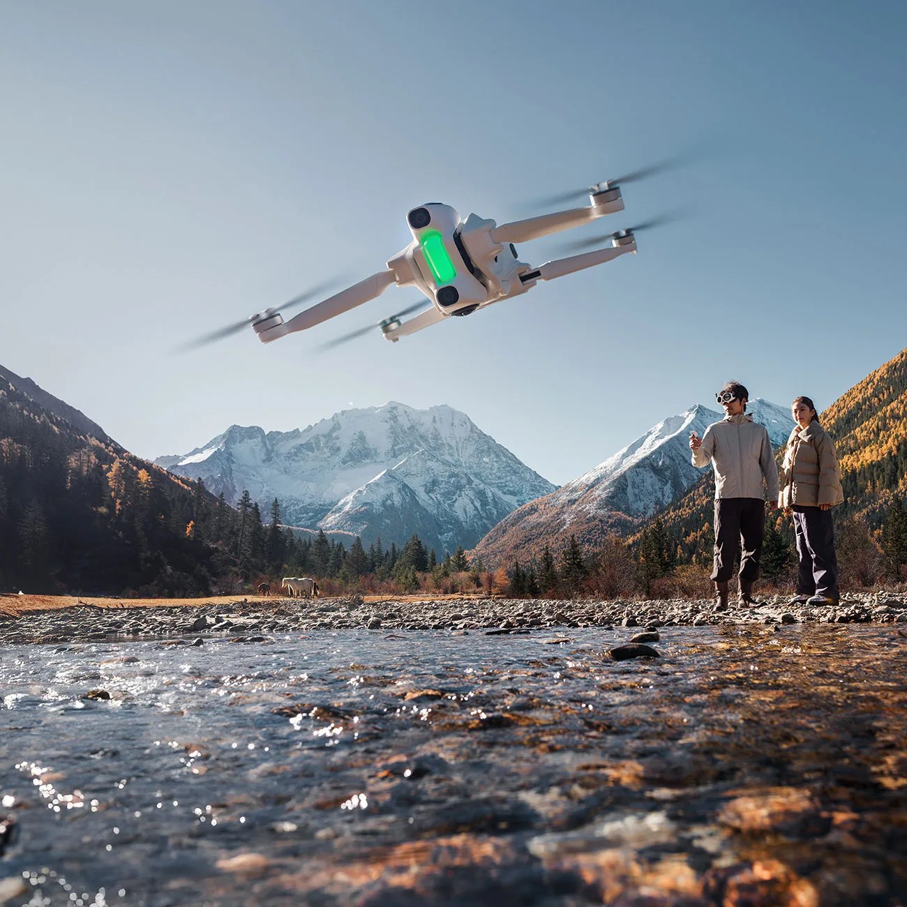 White quadcopter drone with green light flying over a rocky riverbed with two people and snow-capped mountains in the background.