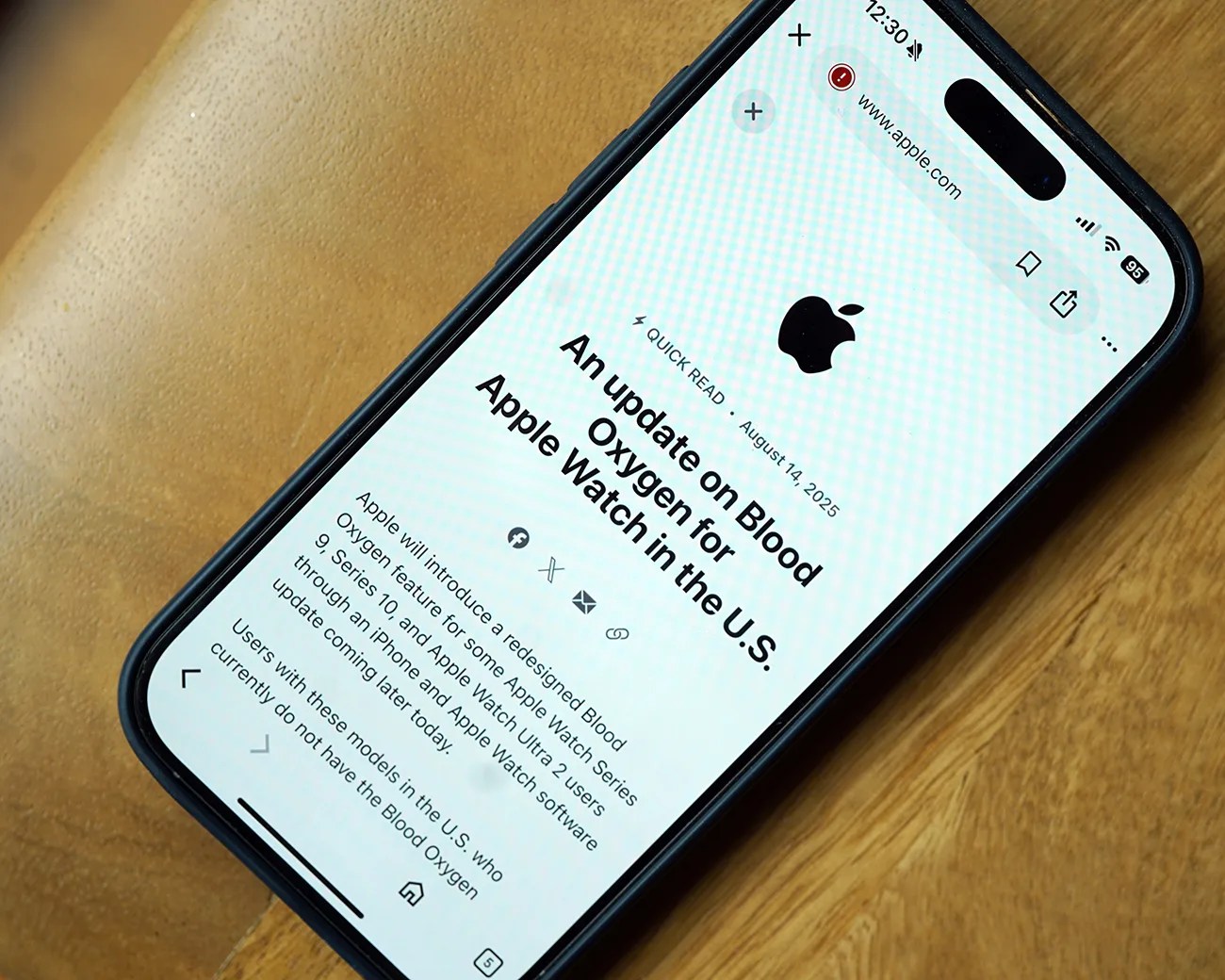 Smartphone displaying an article titled "An update on Blood Oxygen for Apple Watch in the U.S." dated August 14, 2025, with the Apple logo above the title. The article mentions a redesigned Blood Oxygen feature for some Apple Watch Series 9, Series 10, and Apple Watch Ultra 2 users in the U.S., with an update coming later today. The phone is on a wooden surface.