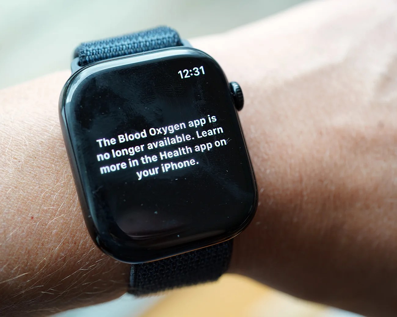 Smartwatch with a black screen displaying a message that reads: "The Blood Oxygen app is no longer available. Learn more in the Health app on your iPhone." The time shown is 12:31. The watch has a dark fabric strap and is worn on a person's wrist.