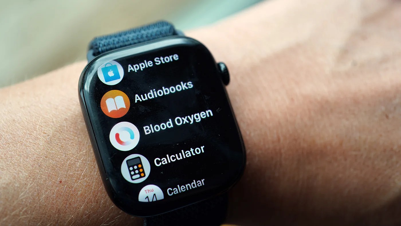 Smartwatch on a wrist displaying app icons and names, including Apple Store, Audiobooks, Blood Oxygen, Calculator, and Calendar. The watch has a dark screen with colorful app icons and a black strap.