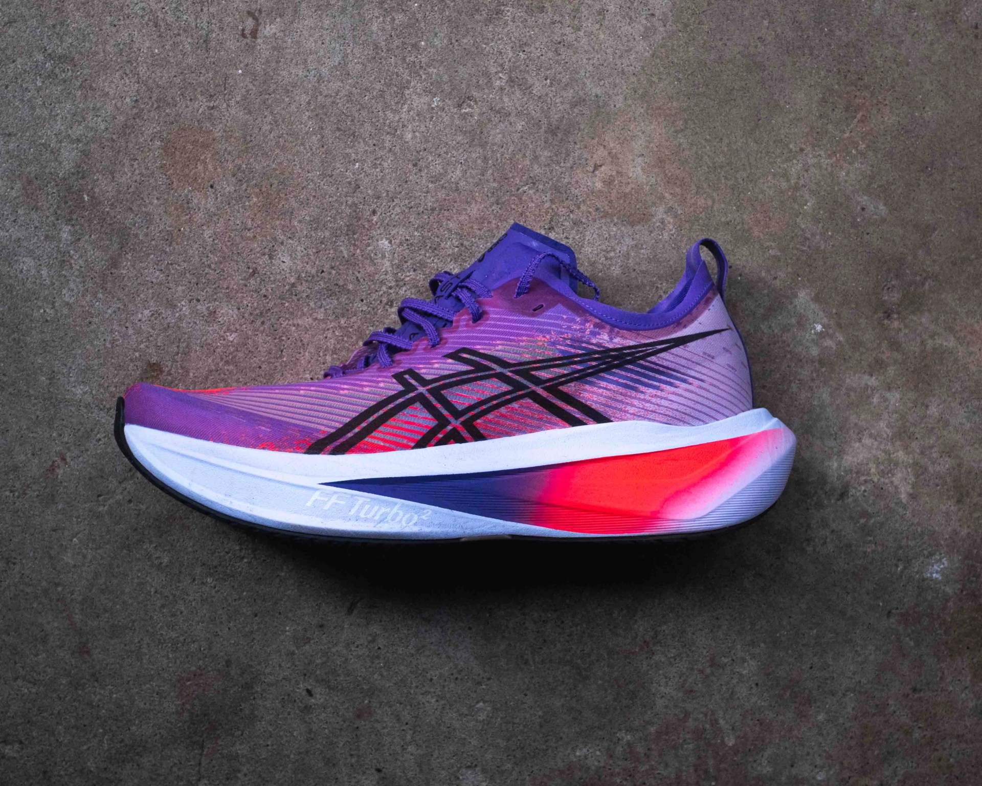 Purple and pink running shoe with black accents and a white sole featuring a red and blue gradient design, placed on a textured concrete surface. The shoe has "FF Turbo²" written on the side of the sole.