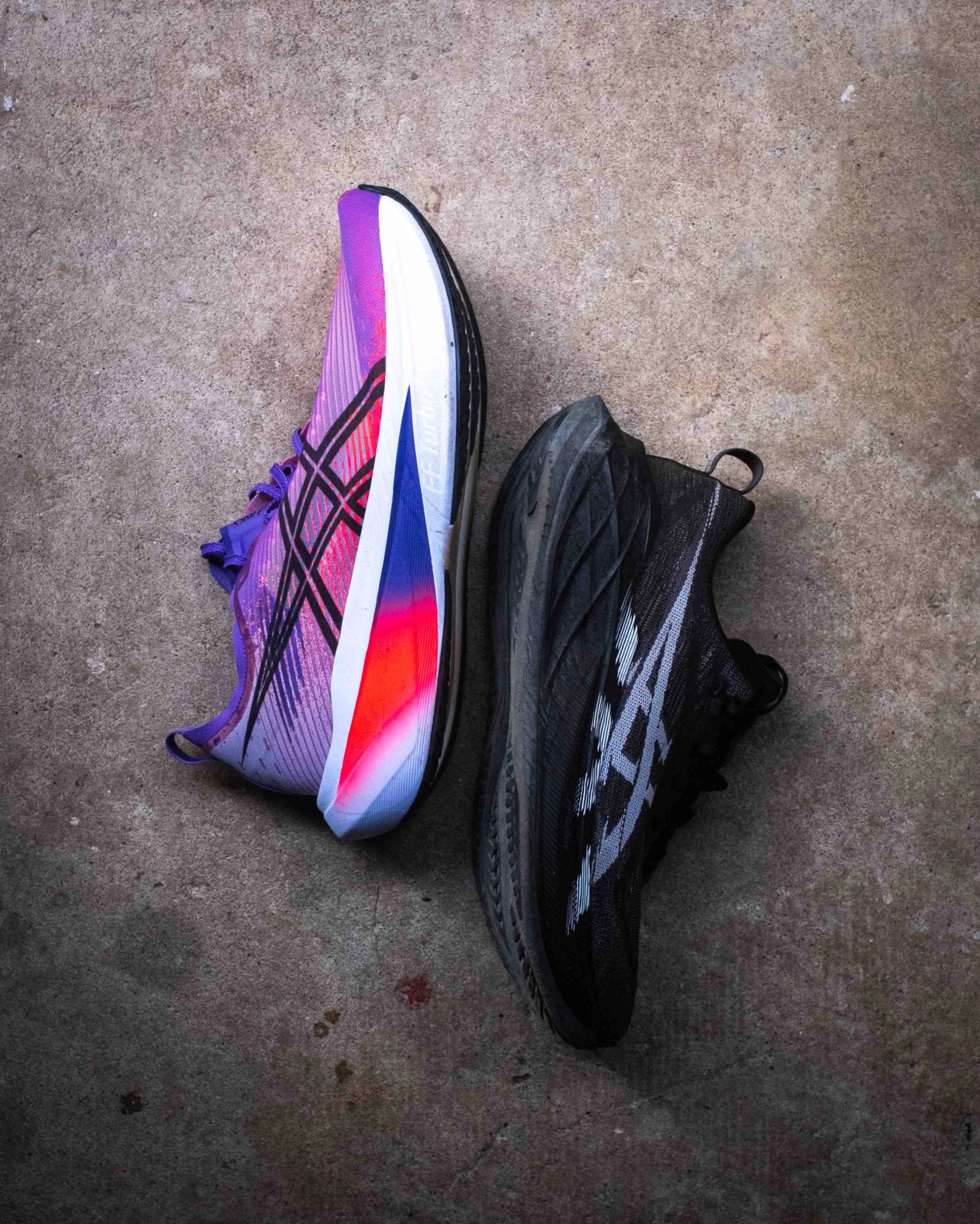 Two athletic shoes placed side by side on a concrete surface: the left shoe is vibrant with a gradient of purple, pink, and red hues with black accents, while the right shoe is predominantly black with white detailing.