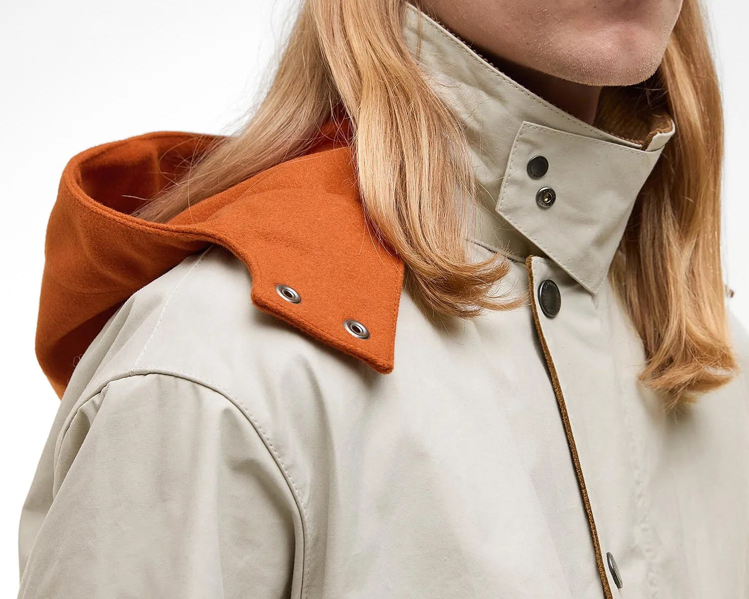 Close-up of a beige jacket with a high collar secured by black snap buttons and an attached burnt orange hood with silver snap fasteners. The jacket is worn by a person with long blonde hair.