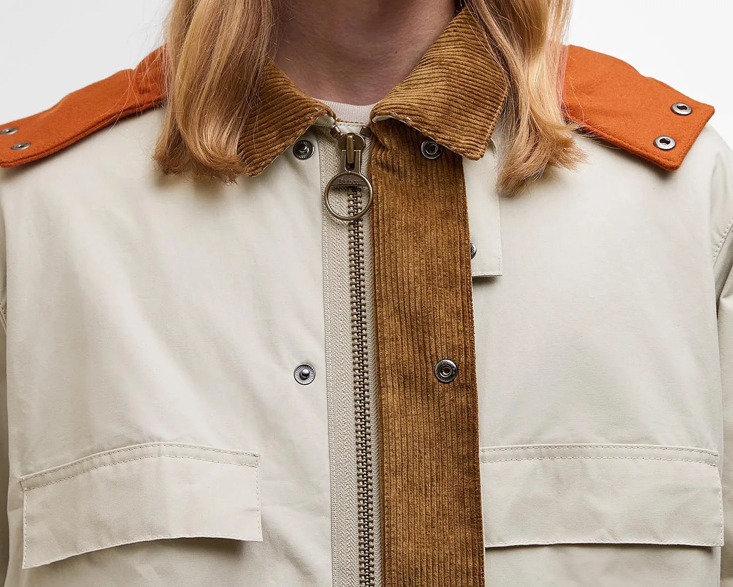 Close-up of a beige jacket featuring a brown corduroy collar and zipper placket. The jacket has silver snap buttons and orange shoulder epaulets with two silver eyelets on each side. The zipper pull is round and metallic. The jacket also includes flap pockets on the chest.