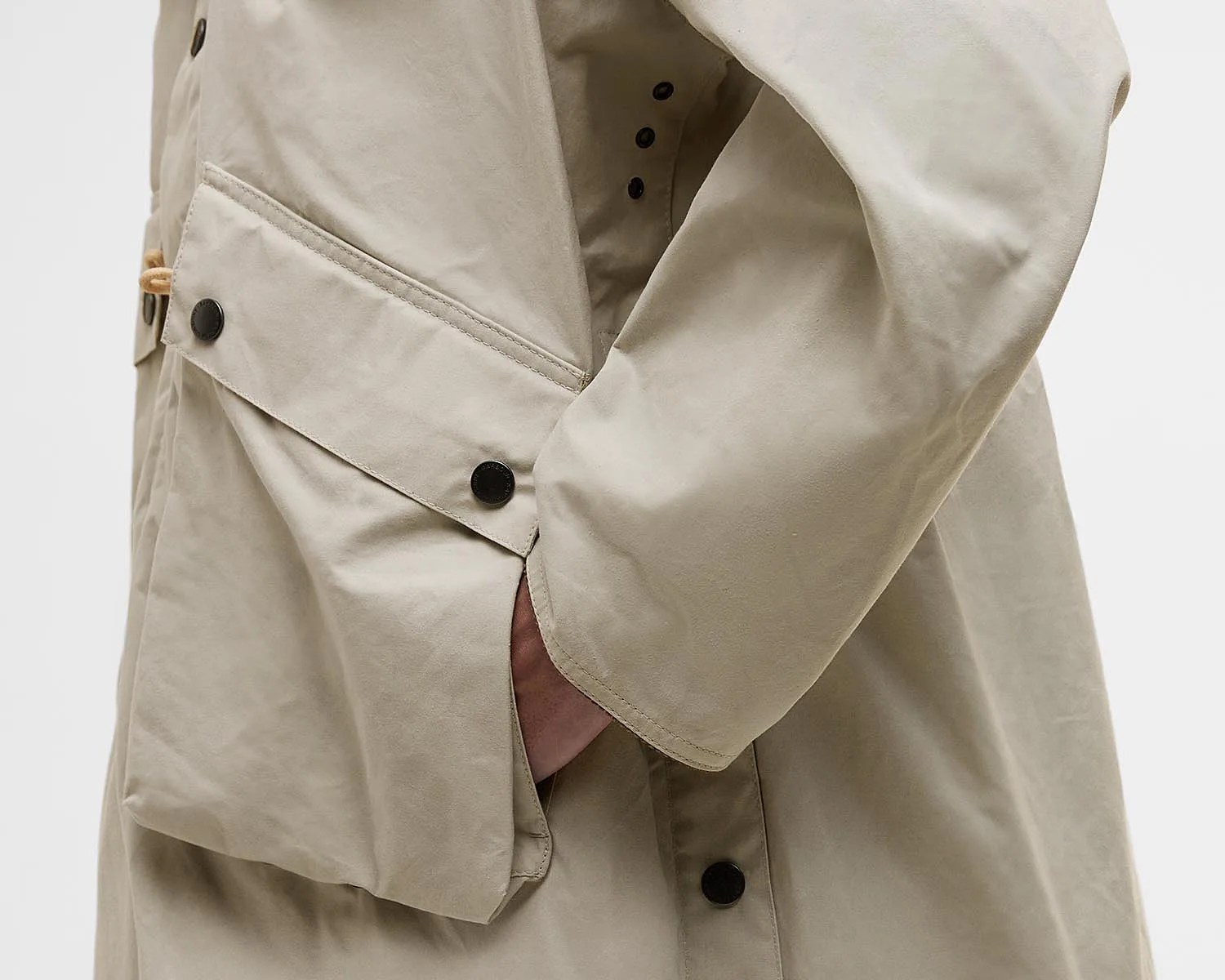 Close-up of a beige parka sleeve and pocket area, featuring black snap buttons and a hand partially tucked into the pocket. The fabric appears smooth and slightly structured.
