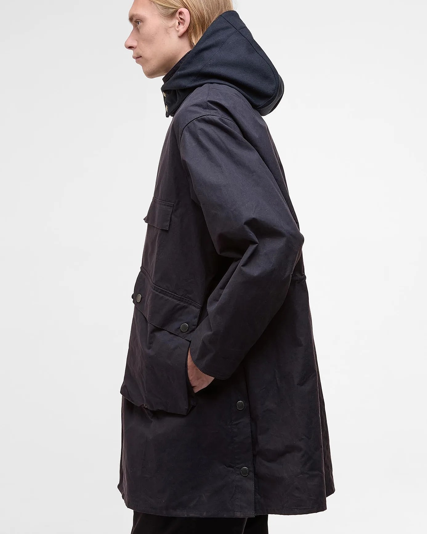 Black long jacket with a hood, featuring large front pockets with buttoned flaps and side snap buttons, worn by a person shown in profile.