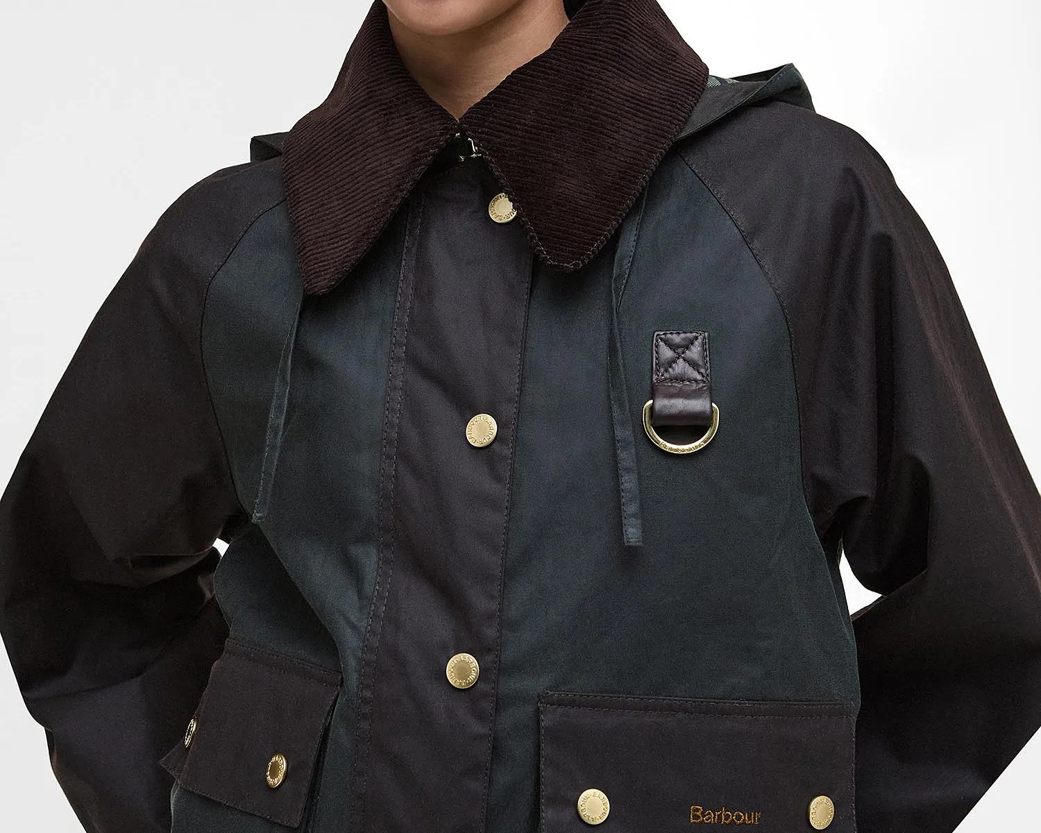 Close-up of a dark-colored Barbour jacket featuring a brown corduroy collar, gold snap buttons down the front, and two front pockets with snap closures. The jacket also has a leather strap with a gold D-ring on the chest.