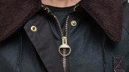 Close-up of a dark green jacket with a brown corduroy collar, featuring a gold zipper pull with the brand name "Barbour" and gold snap buttons on the front flap.