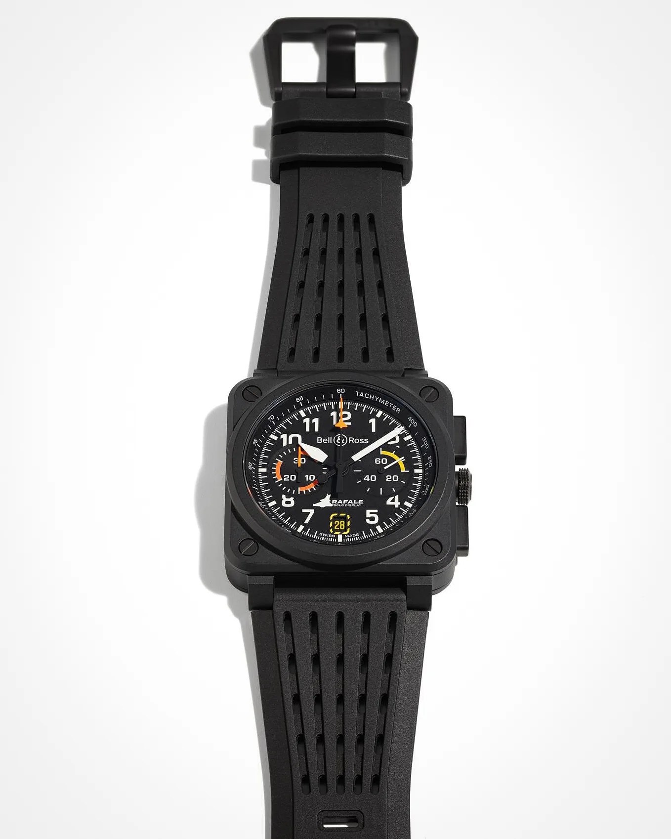 Black Bell & Ross wristwatch with a square case and a black perforated rubber strap. The watch face features white numerals, orange and yellow accents on the subdials, and a date window at the 6 o'clock position. The dial includes the text "Bell & Ross," "Rafale," and "Swiss Made," with a tachymeter scale around the outer edge.