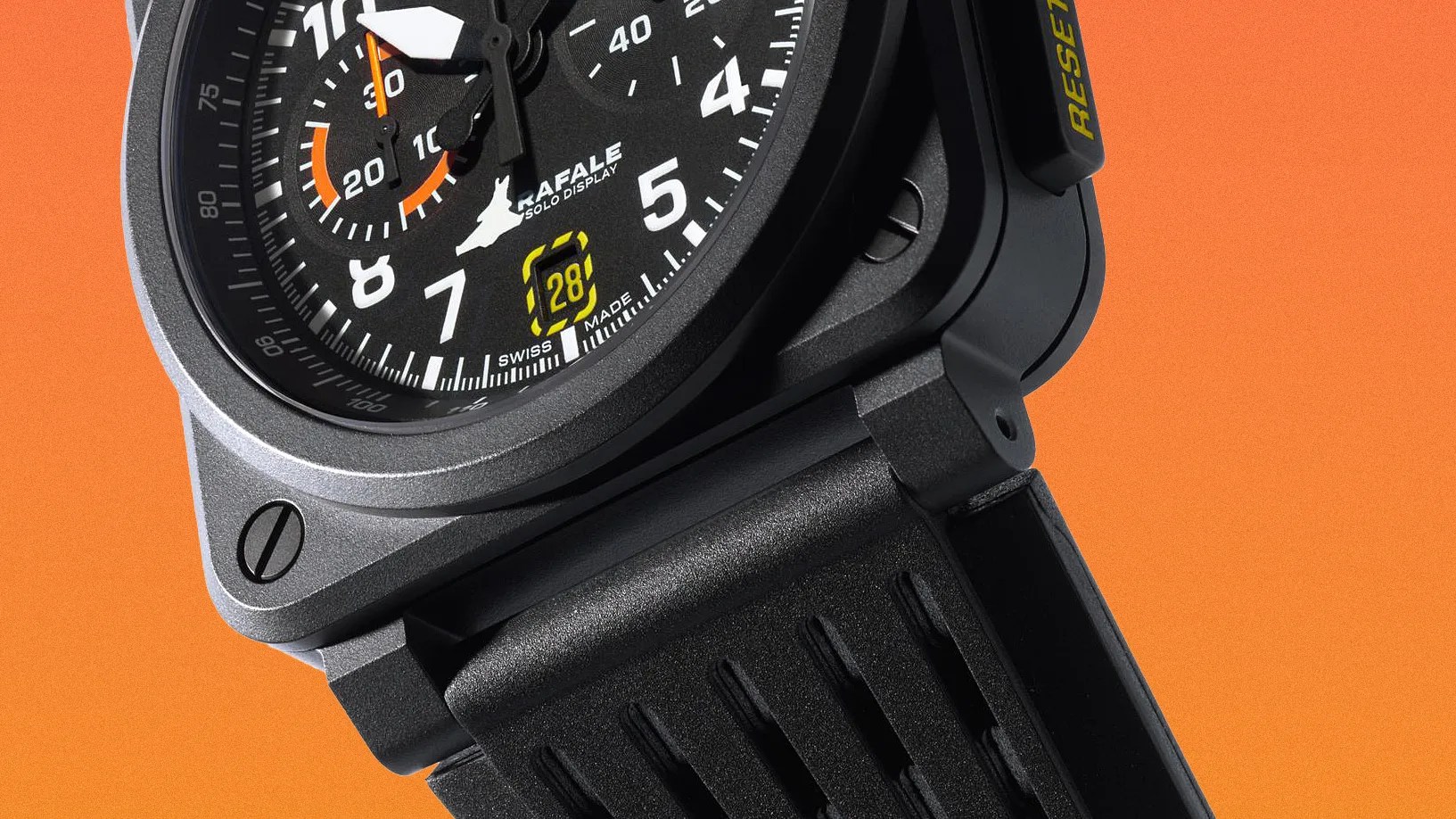 Close-up of a black aviation-style wristwatch with a round dial featuring white numerals and markings, orange accents on the sub-dial and second hand, and a date window displaying "28" in yellow. The watch face includes the text "RAFALE SOLO DISPLAY" and "SWISS MADE." The watch has a robust black strap and a "RESET" button on the side with yellow lettering, set against an orange background.