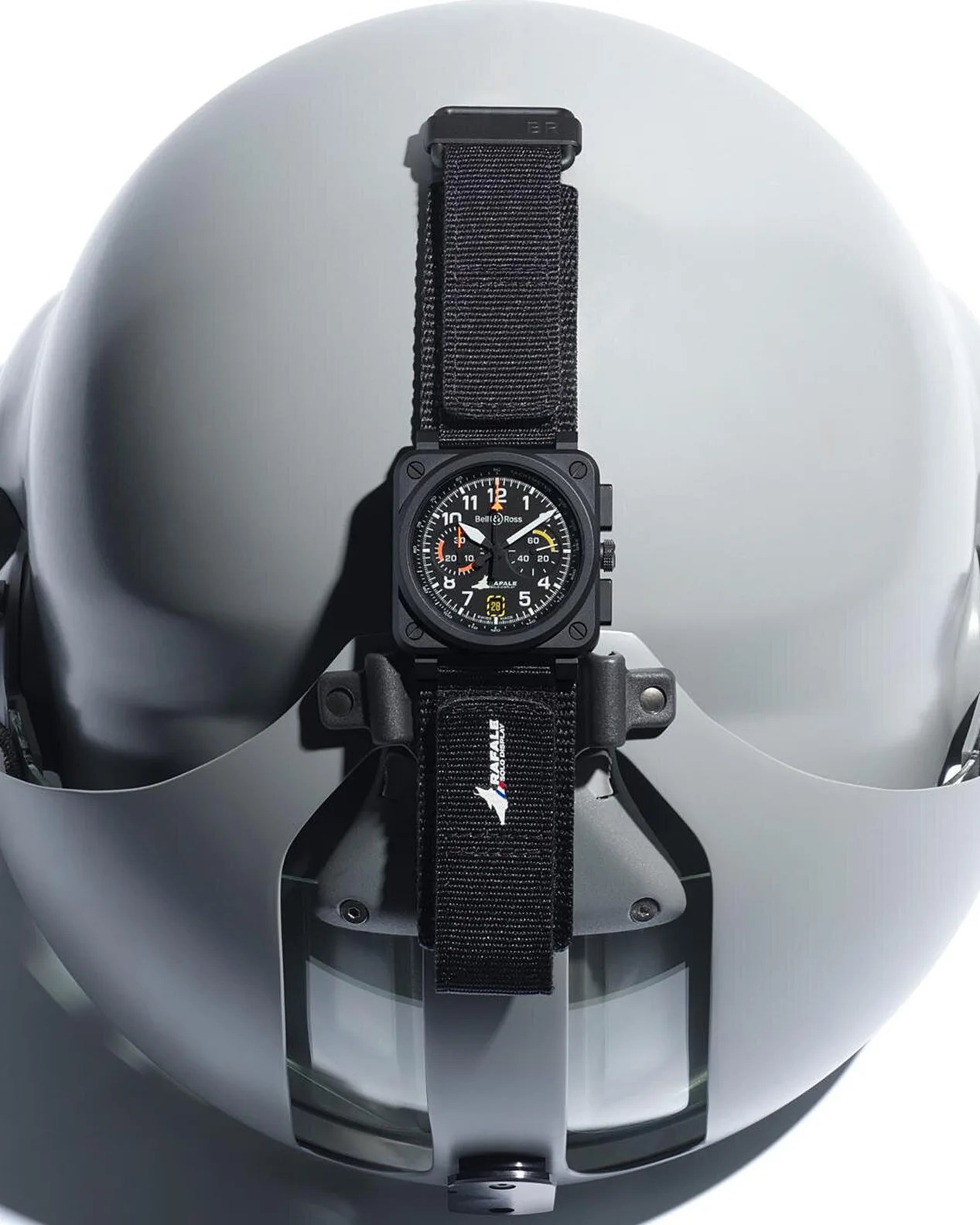 A black Bell & Ross wristwatch with a square case and a black fabric strap is mounted on a light gray pilot helmet. The watch face features white numerals, orange and yellow accents, and multiple subdials.