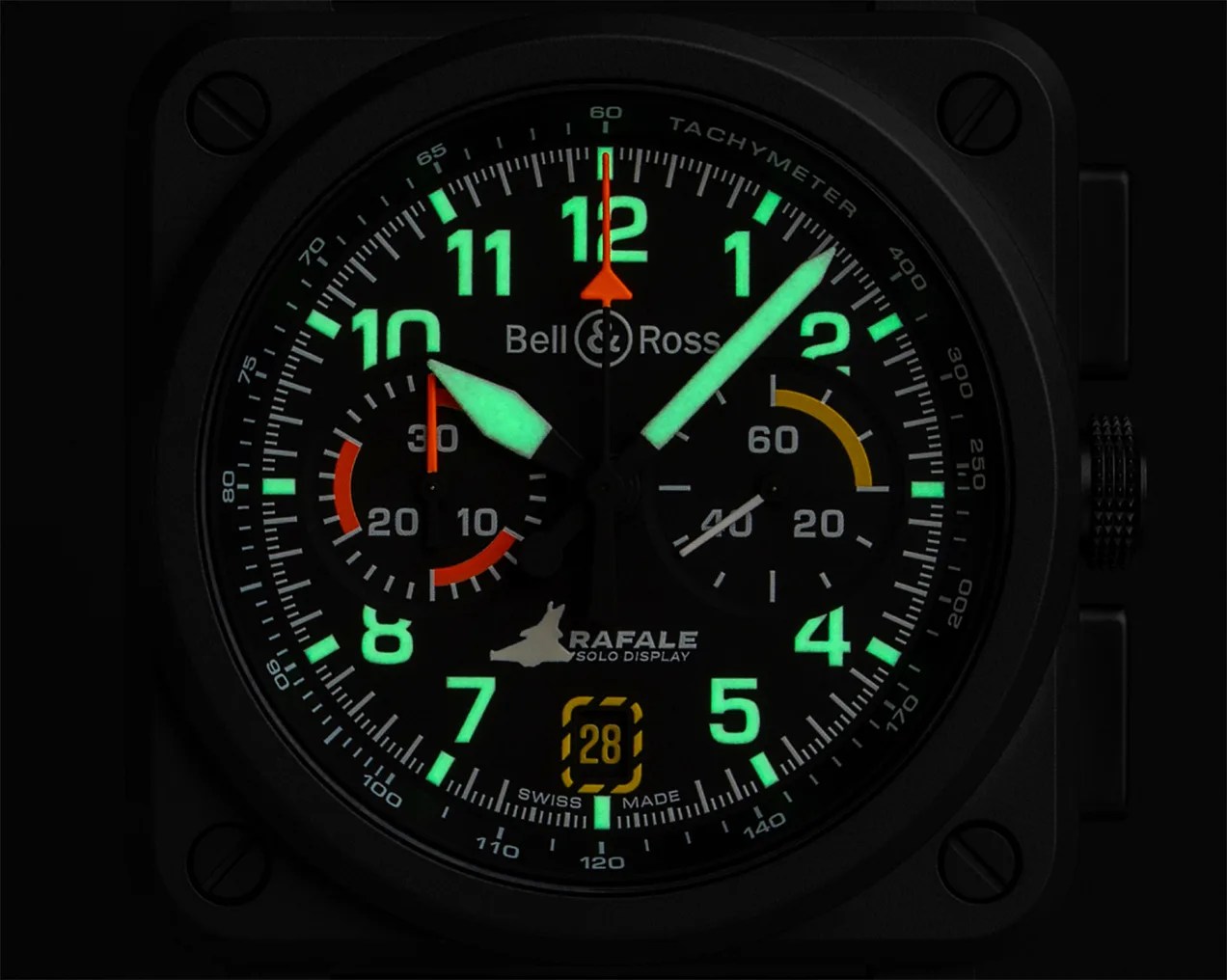 Square black Bell & Ross wristwatch with luminous green Arabic numerals and hands. The watch face features two subdials with orange and yellow accents, a date window at the 6 o'clock position displaying "28," and the text "RAFALE SOLO DISPLAY" above the date. The bezel has a tachymeter scale, and the watch has a black crown and pushers on the right side.