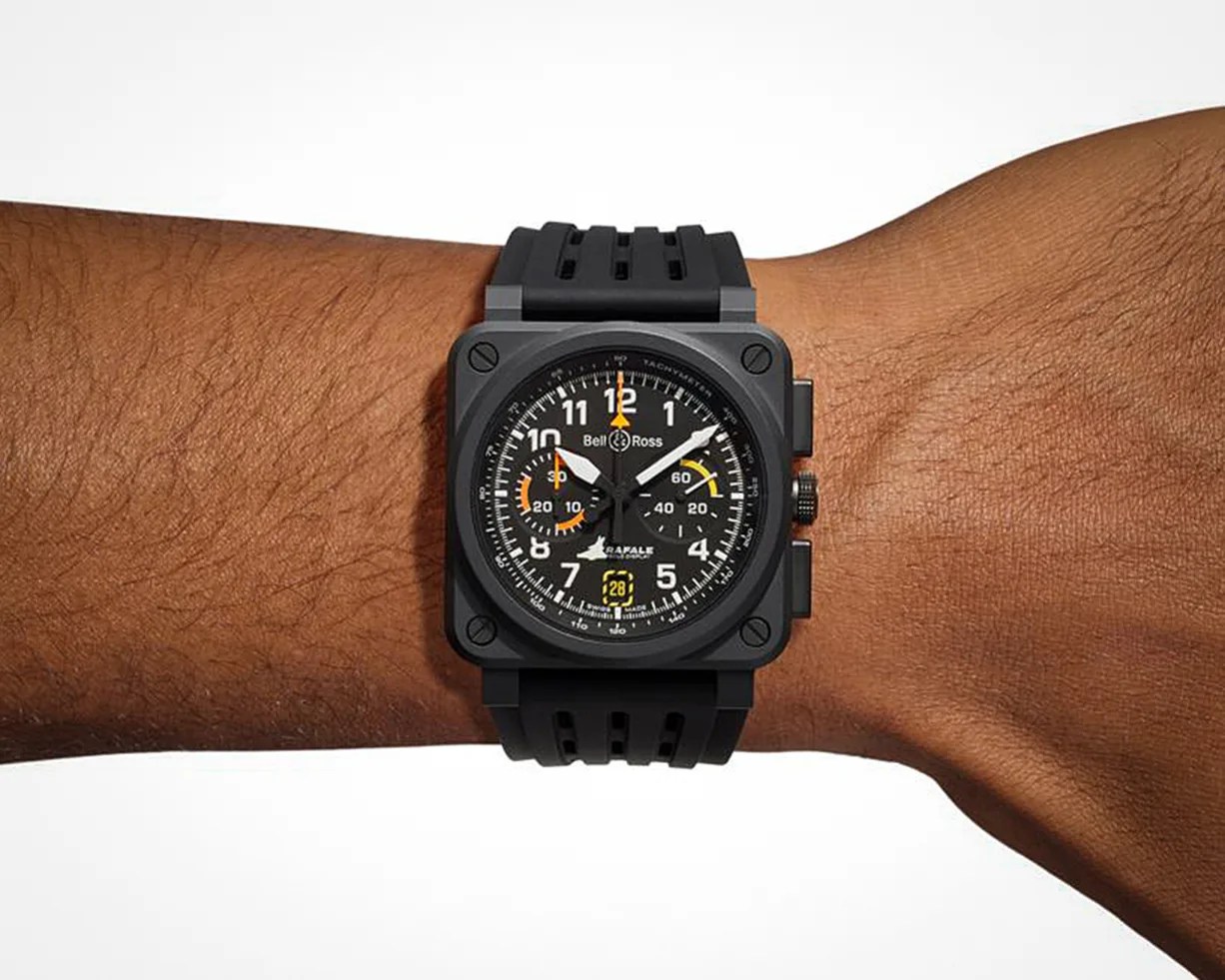 Black Bell & Ross wristwatch with a square case and round dial, featuring white numerals and hands, orange and yellow accents, and a black rubber strap, worn on a person's wrist.