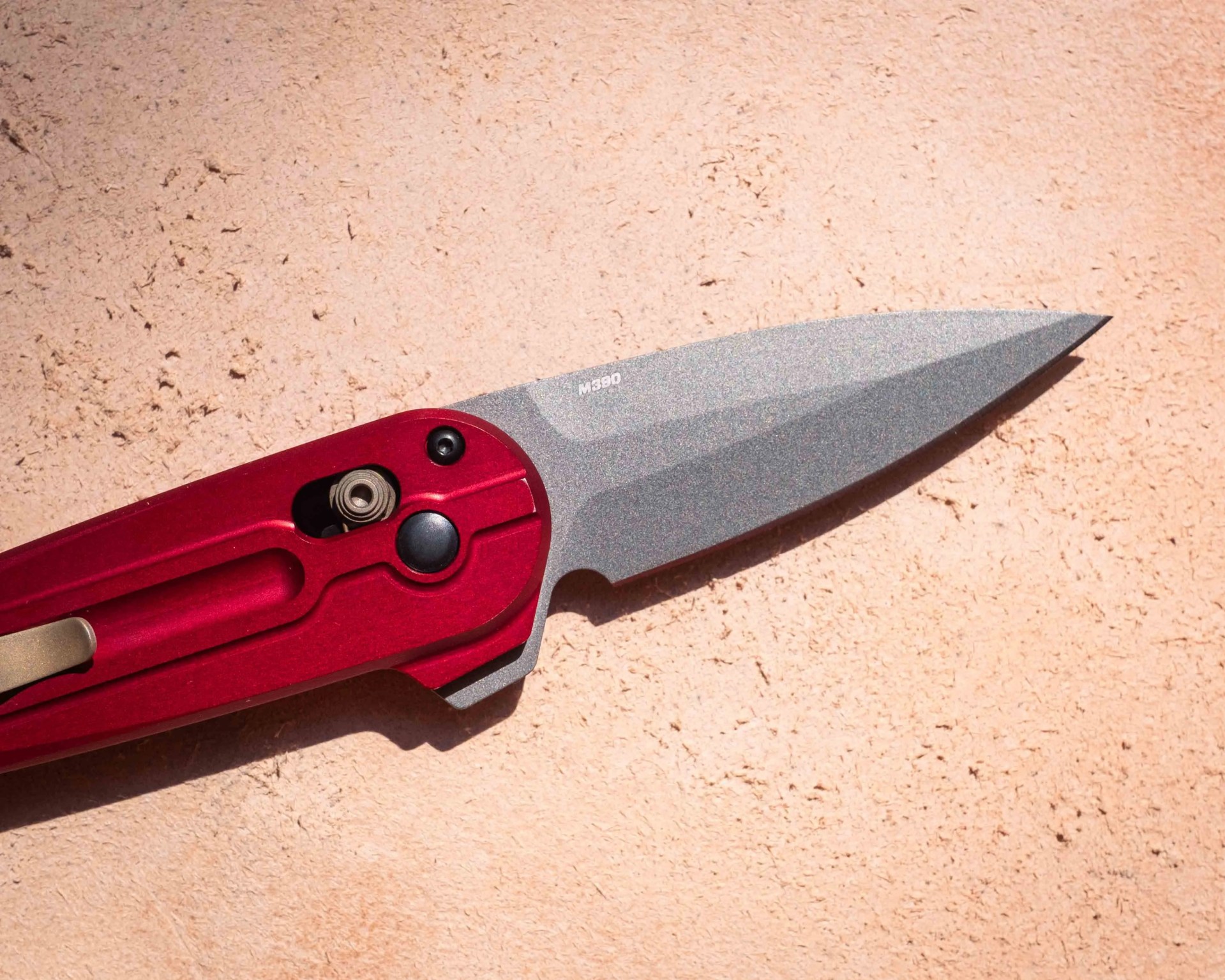 Folding knife with a red handle and a gray stonewashed blade marked "M390," placed on a textured light brown surface.