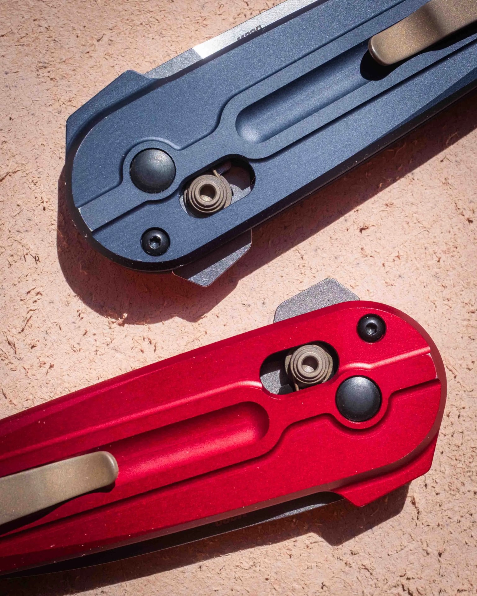 Two closed folding knives with metal handles, one in matte blue and the other in matte red, placed on a textured light brown surface. Both knives feature a visible pivot mechanism and a pocket clip.