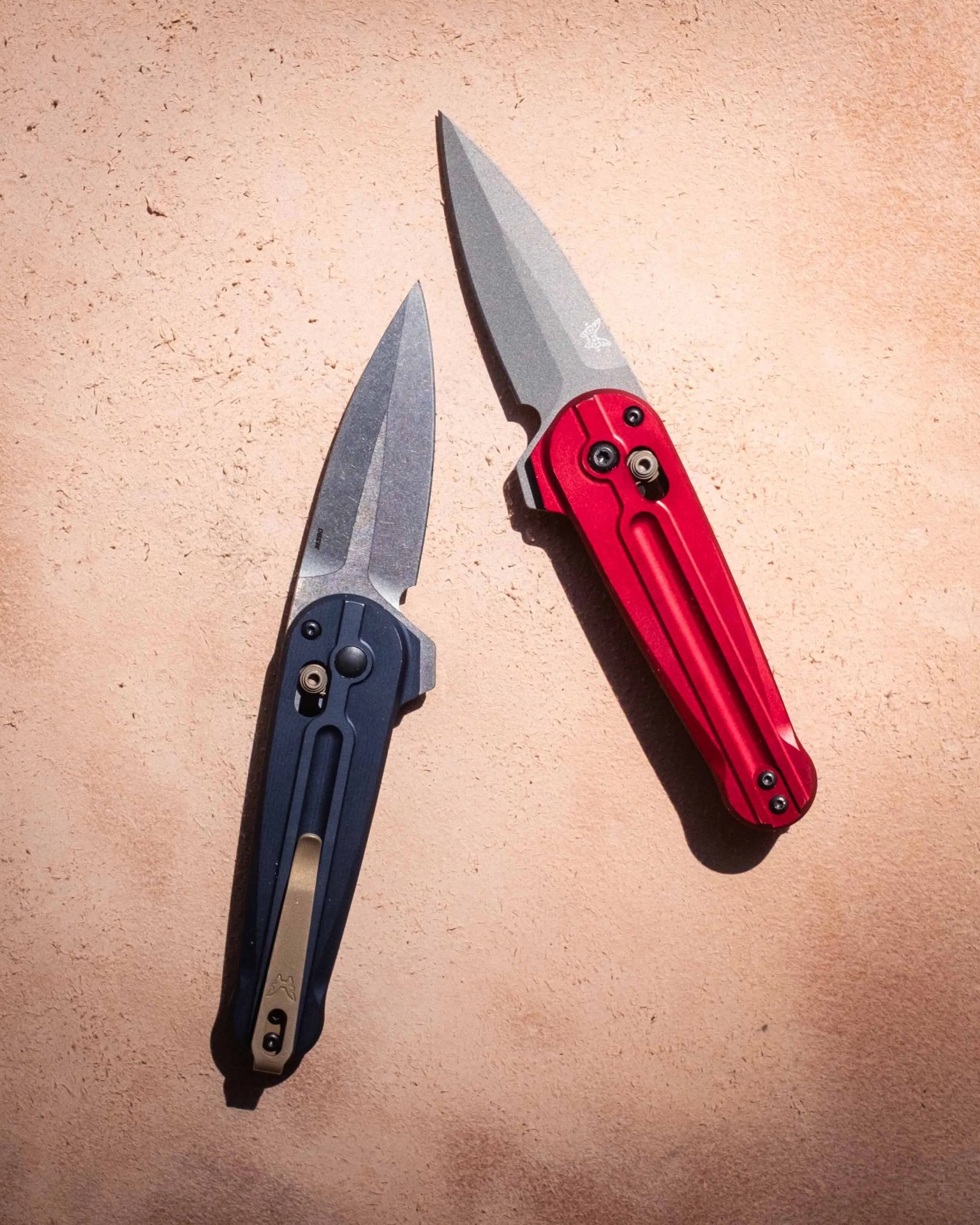 Two folding knives with stonewashed blades are shown on a textured surface. One knife has a dark blue handle with a bronze pocket clip, while the other has a red handle with black screws and hardware. Both knives have a similar blade shape and locking mechanism.