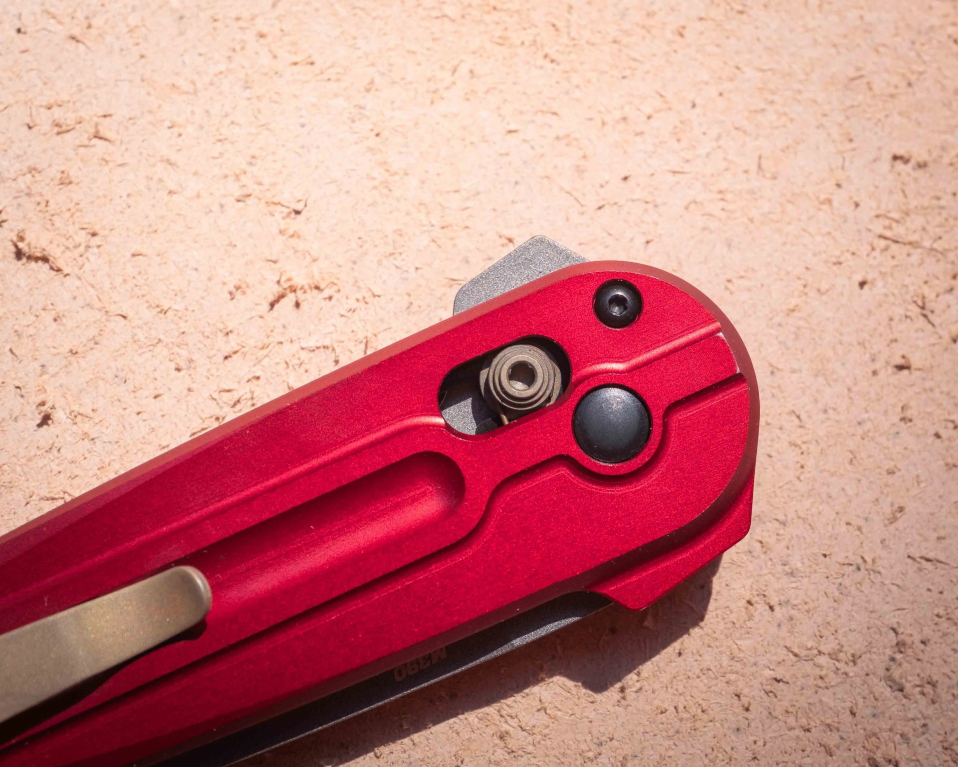 Close-up of the handle end of a red folding knife with a visible pivot mechanism and a black button or screw on a textured beige surface.