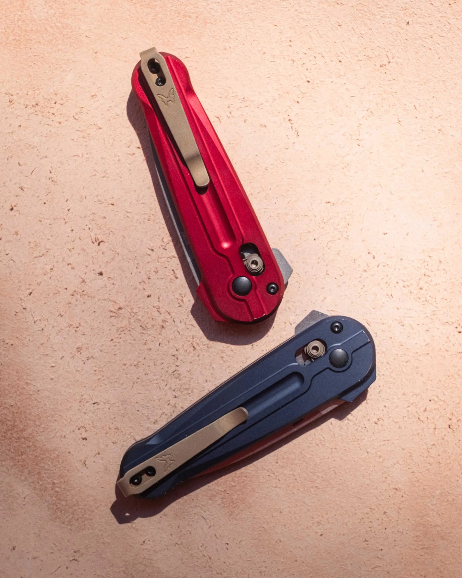 Two closed folding knives with metal clips are shown on a textured beige surface. One knife has a red handle with a bronze-colored clip, and the other has a dark blue handle with a similar bronze-colored clip. Both clips feature a small butterfly logo. The knives have visible pivot screws and locking mechanisms near the base of the handles.