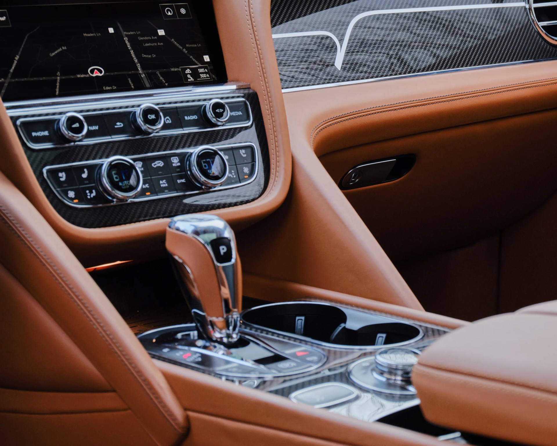 Car interior center console featuring a gear shift lever with a "P" button, dual cup holders, climate control knobs, media and navigation buttons, and a touchscreen displaying a navigation map. The interior is upholstered in brown leather with carbon fiber trim accents.