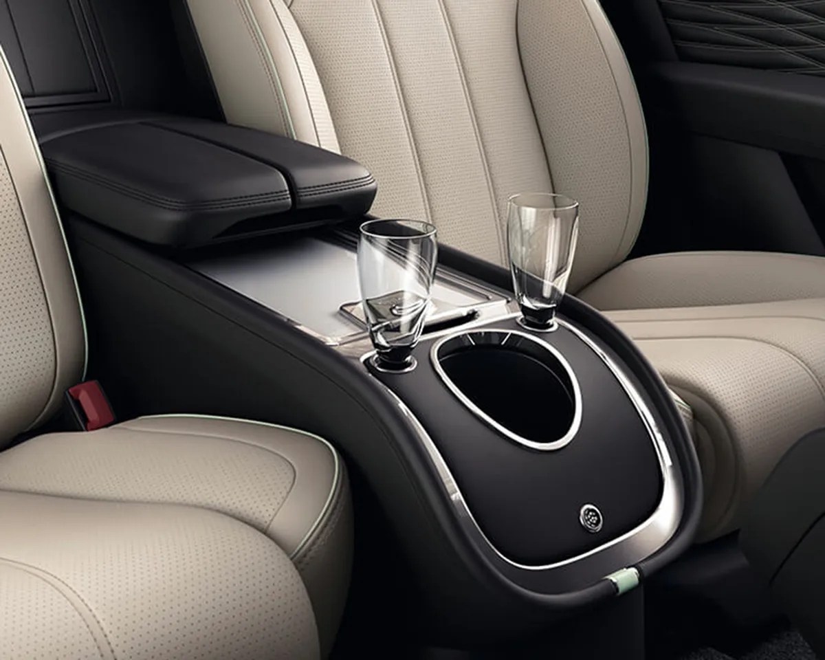 Car interior center console with beige perforated leather seats, featuring a black armrest, two transparent glasses in cup holders, and a sleek black and chrome trim design.