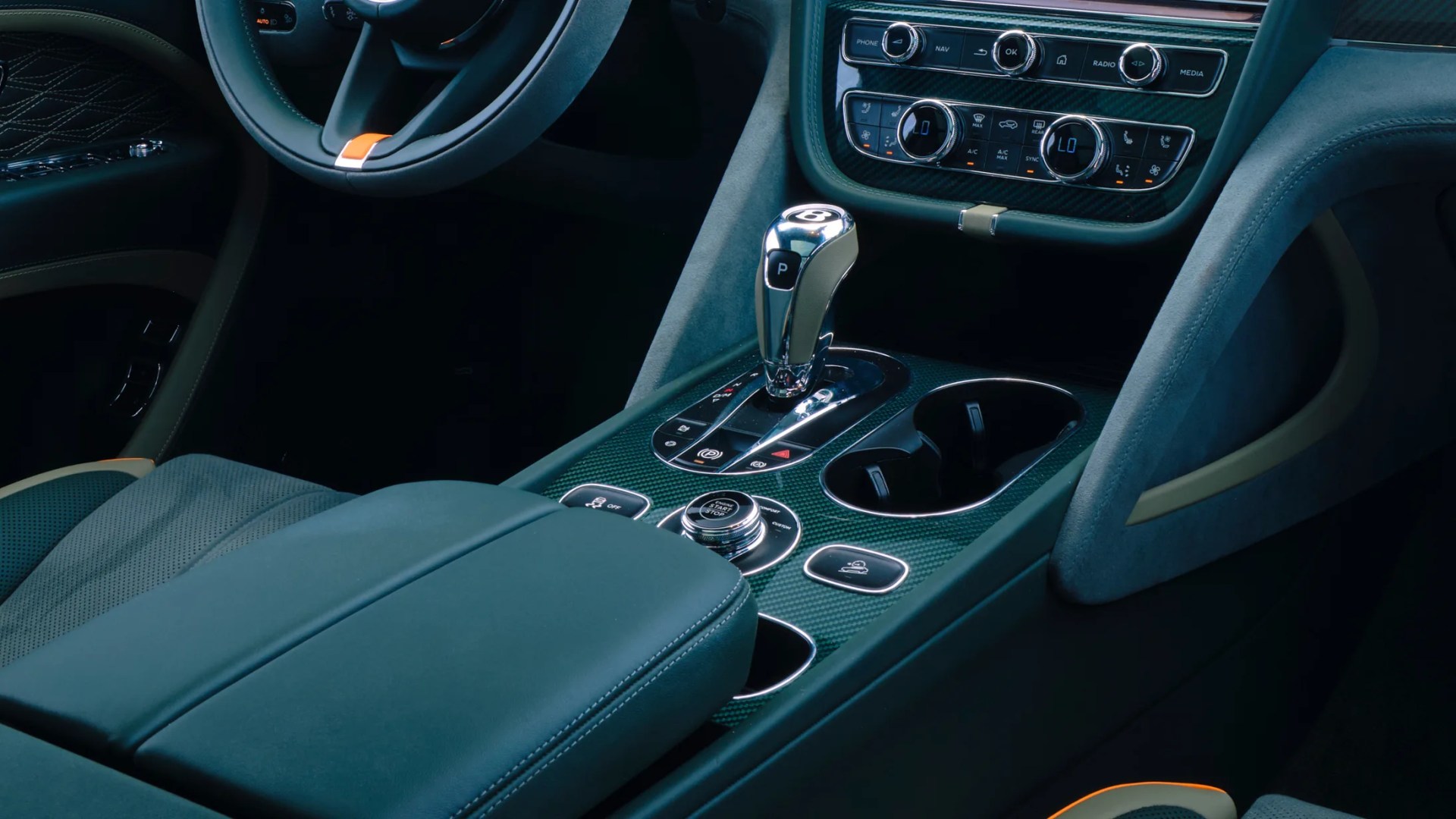 Center console of a luxury car featuring a chrome gear shifter with the Bentley logo, surrounded by carbon fiber trim. The console includes cup holders, various control buttons, and a rotary dial for engine start/stop. The interior is upholstered in dark green leather with orange accents and detailed stitching. The dashboard above shows climate control and media buttons.