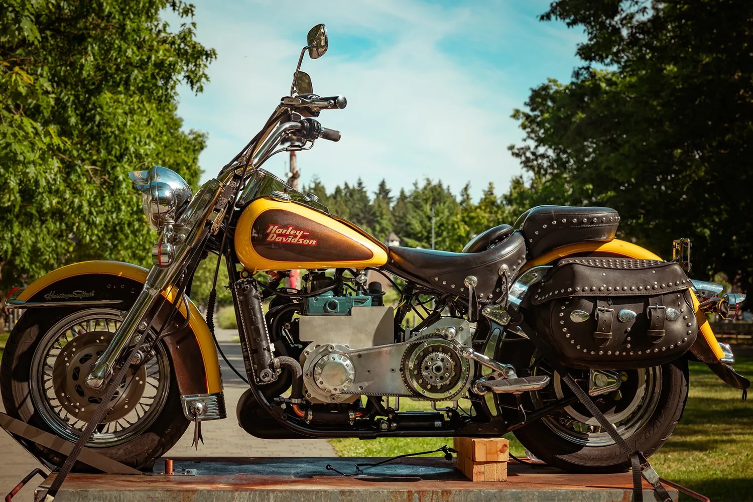 Yellow and black Harley-Davidson motorcycle with leather studded saddlebags, displayed outdoors on a platform with trees and a blue sky in the background.