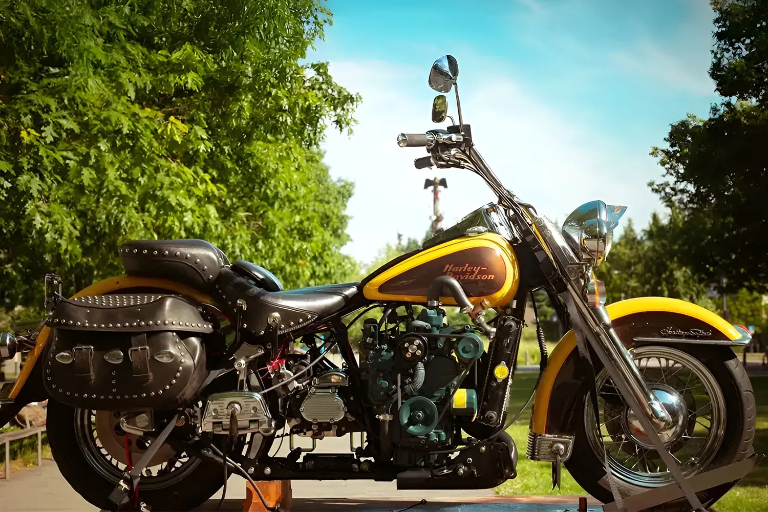 Vintage Harley-Davidson motorcycle with a yellow and brown fuel tank, black leather seat, and studded black leather saddlebags, parked outdoors with green trees and a clear blue sky in the background.