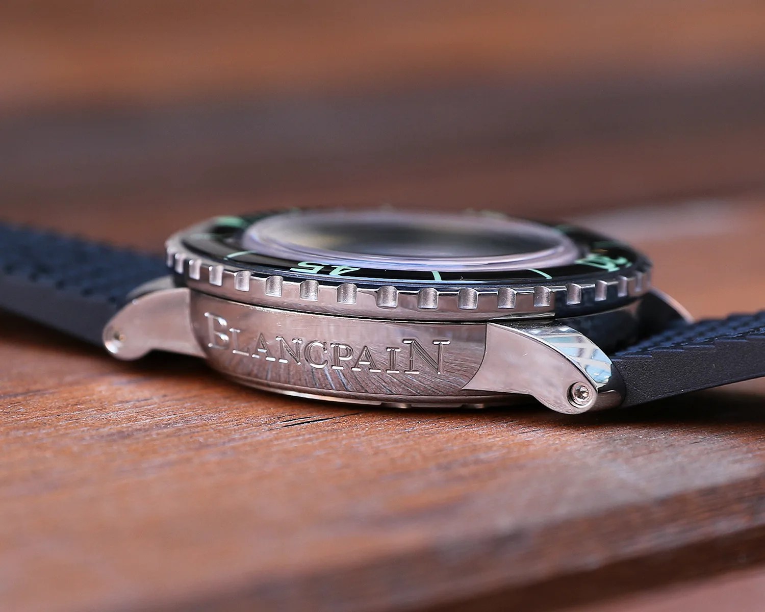 Close-up side view of a Blancpain wristwatch with a black bezel featuring green markings, a domed crystal, and a black textured strap. The polished metal case has "BLANCPAIN" engraved on the side. The watch rests on a wooden surface.