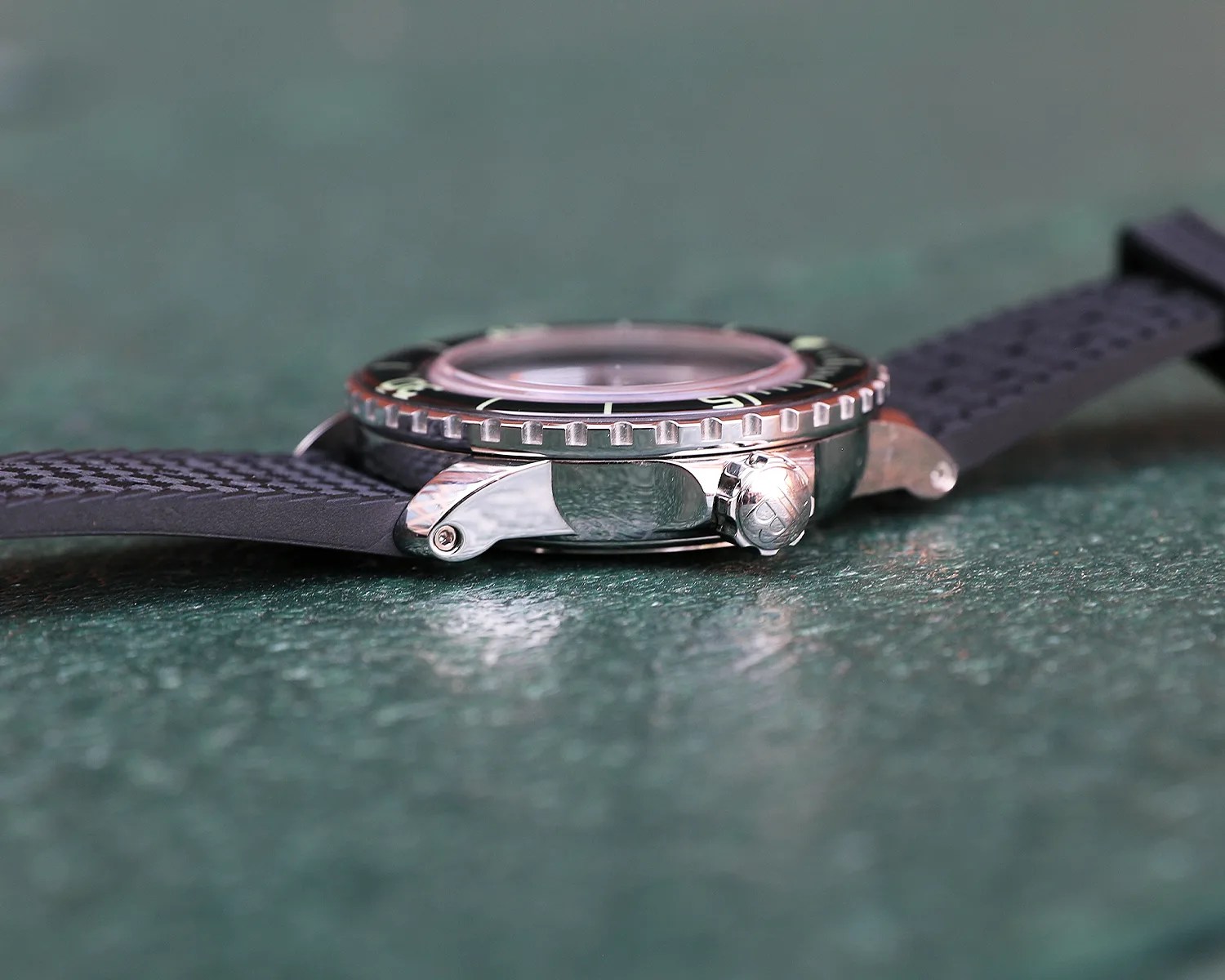 Side view of a blancpain wristwatch with a black textured rubber strap, a polished stainless steel case, and a ridged bezel. The crown is visible on the right side of the case, featuring an engraved logo. The watch is resting on a green textured surface.