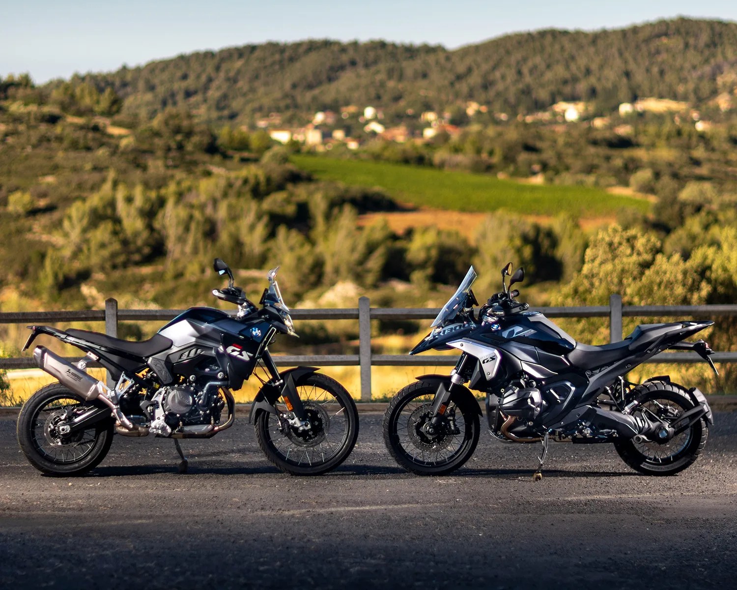 Two black and silver BMW adventure motorcycles are parked side by side on a paved surface with a wooden fence and green hills in the background. Both bikes have windshields and off-road tires, with the one on the right appearing slightly larger and more robust.