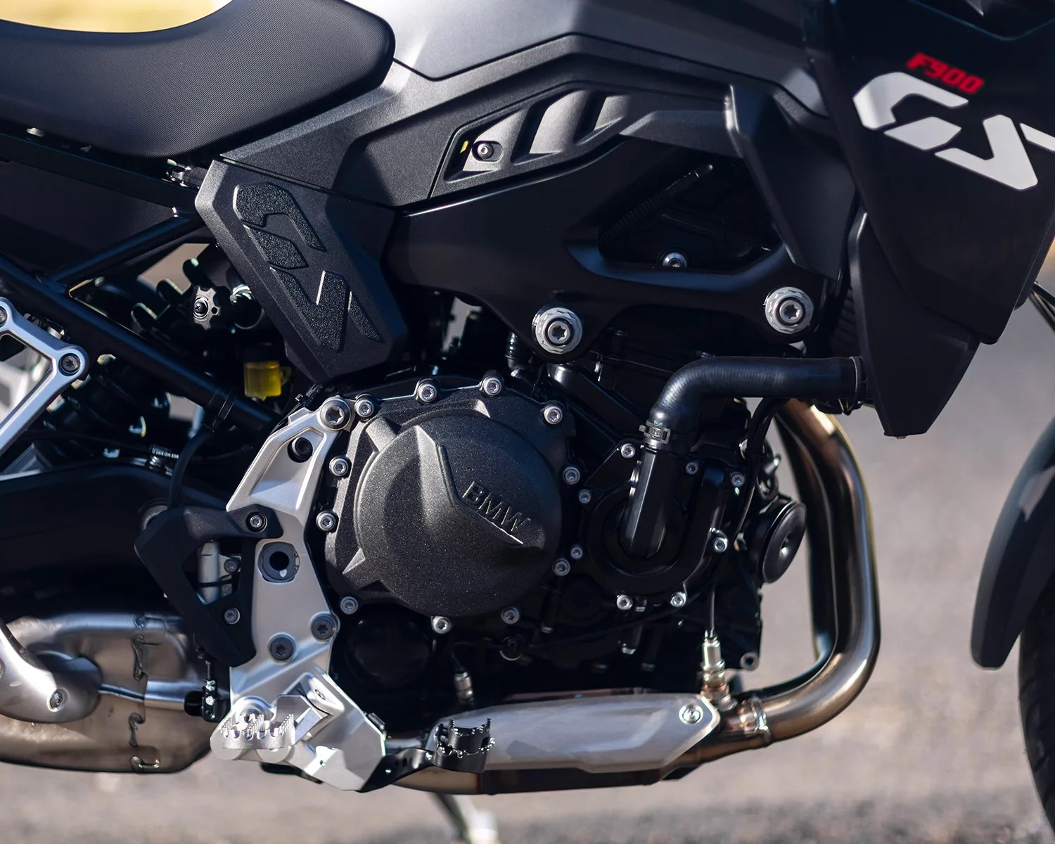 Close-up of a BMW motorcycle engine, showing the black engine cover with the BMW logo, surrounding bolts, a black hose, and part of the exhaust pipe. The frame and footpeg are also visible, along with the "F900" and "GX" markings on the black body panel above the engine.