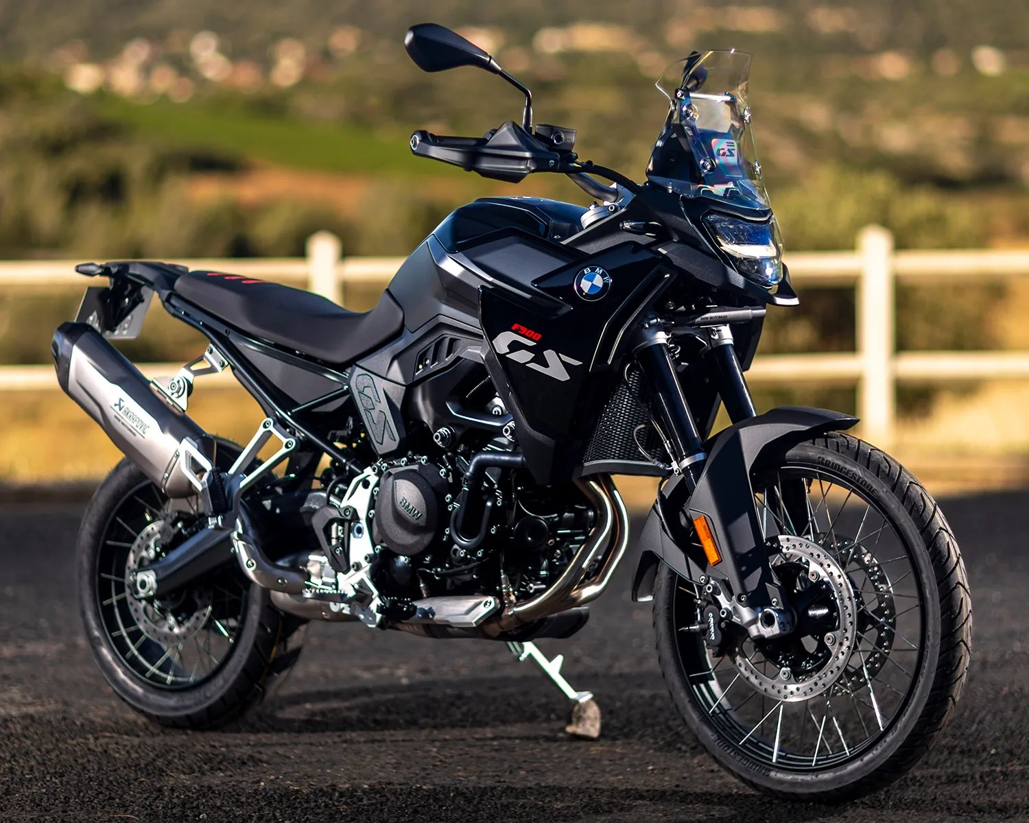 Black BMW F 900 GS adventure motorcycle with a clear windshield, spoked wheels, and an Akrapovič exhaust, parked on a paved surface with a blurred natural background.
