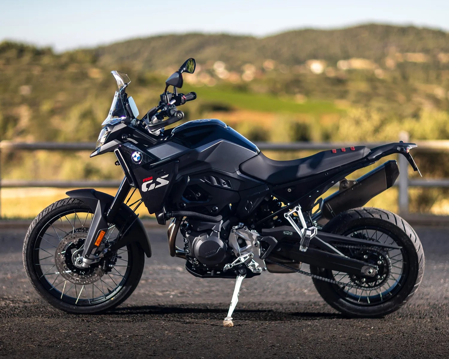 Black BMW F 900 GS adventure motorcycle parked on a paved surface with a blurred natural landscape in the background. The bike features a clear windshield, spoked front wheel, and a large exhaust on the right side.